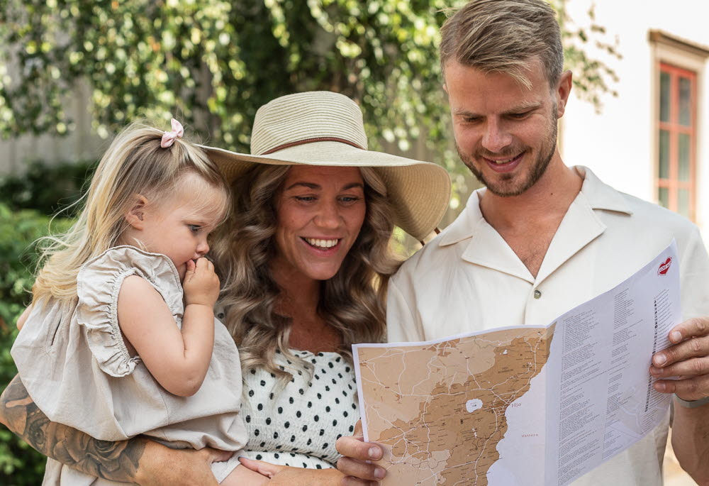 
A woman in a polka dot dress and a straw hat is standing, holding her daughter, and a man next to her is holding a map and they are looking at it.