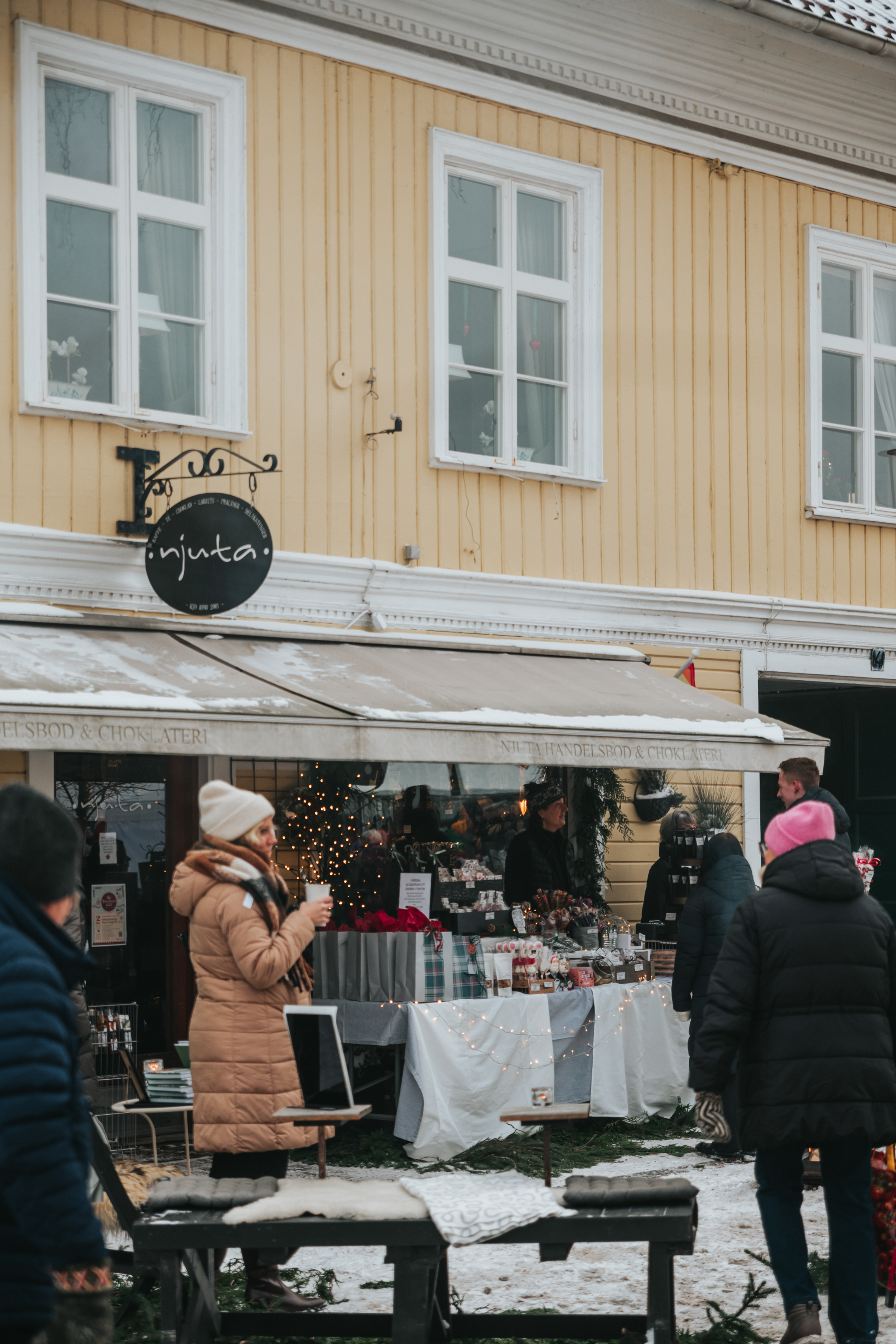 Människor står utanför en butik, huset är i trä och gult. En svart skylt där det står njuta hänger på väggen. Det är snö på marken. 