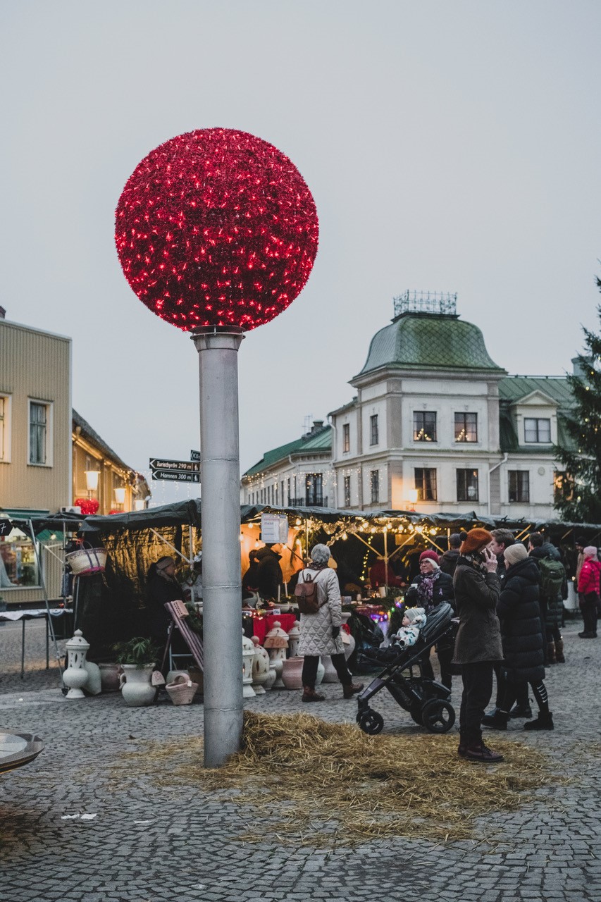 En stolpe med en röd boll på på ett torg. Folk runt omkring. 