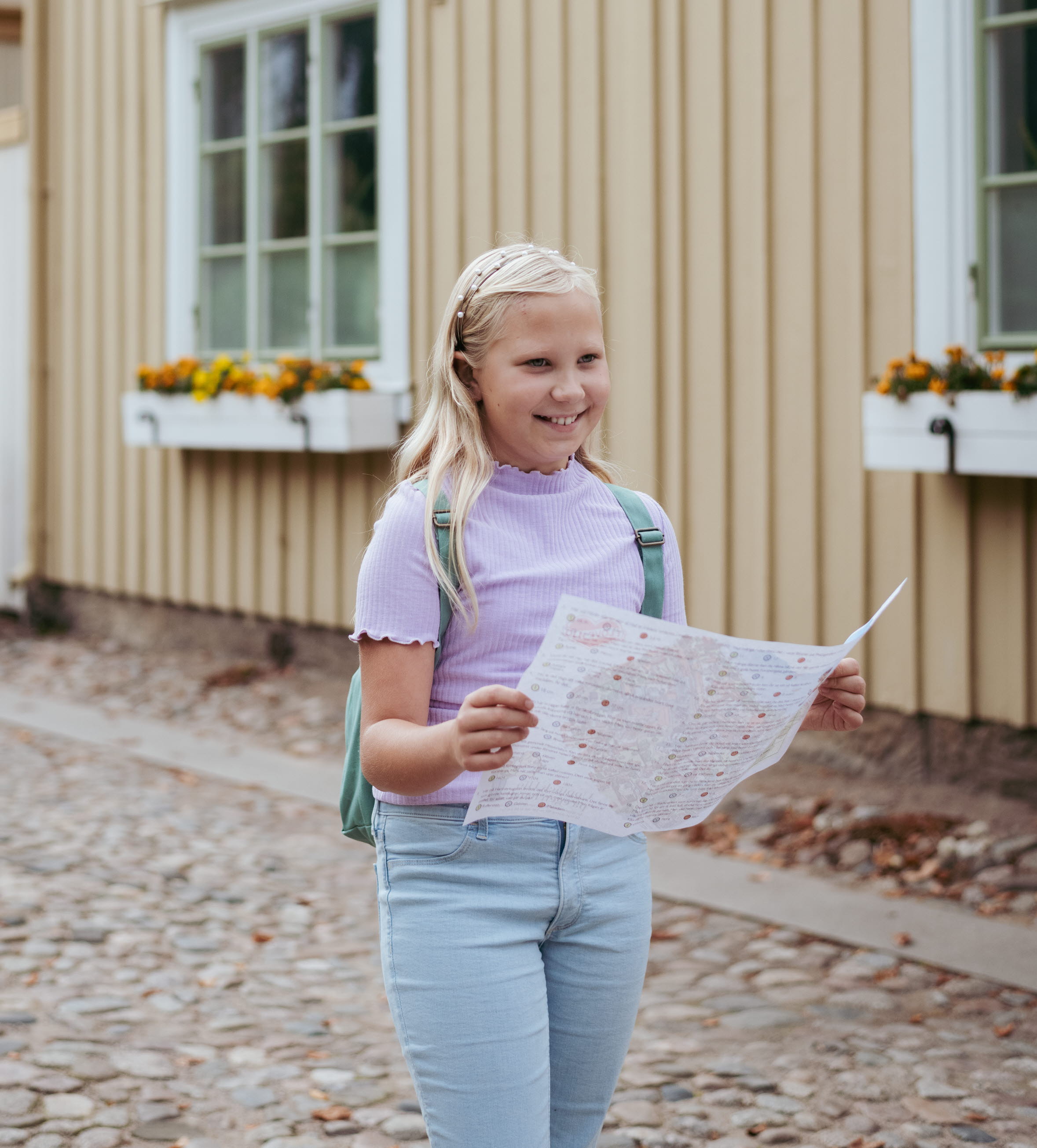 Flicka med ljus långt hår som har en turkos ryggsäck, jeans och en lila tröja håller i en karta. Gult trähus i bakgrunden