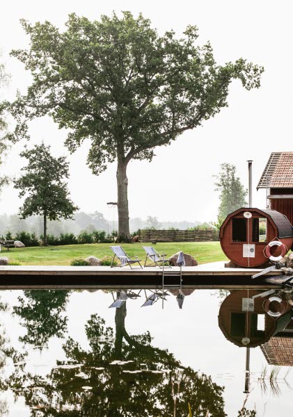 Vatten i förgrunden, med två solstolar på däck och en rund liten röd bastu bredvid. Det är ett stort träd bakom som speglar sig i vattnet. 