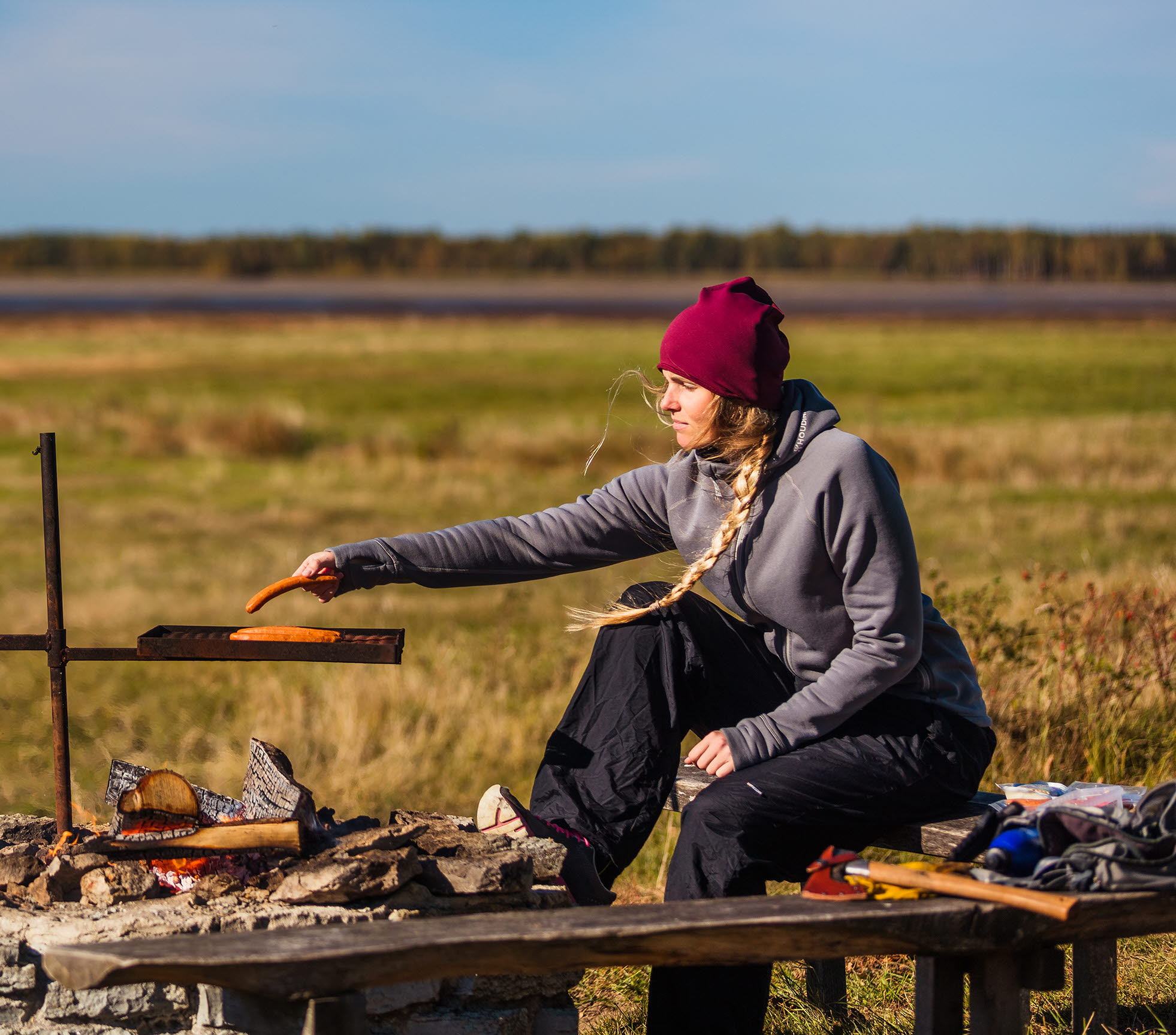 Kvinna som sitter vid en grillplats och grillar en korv. 