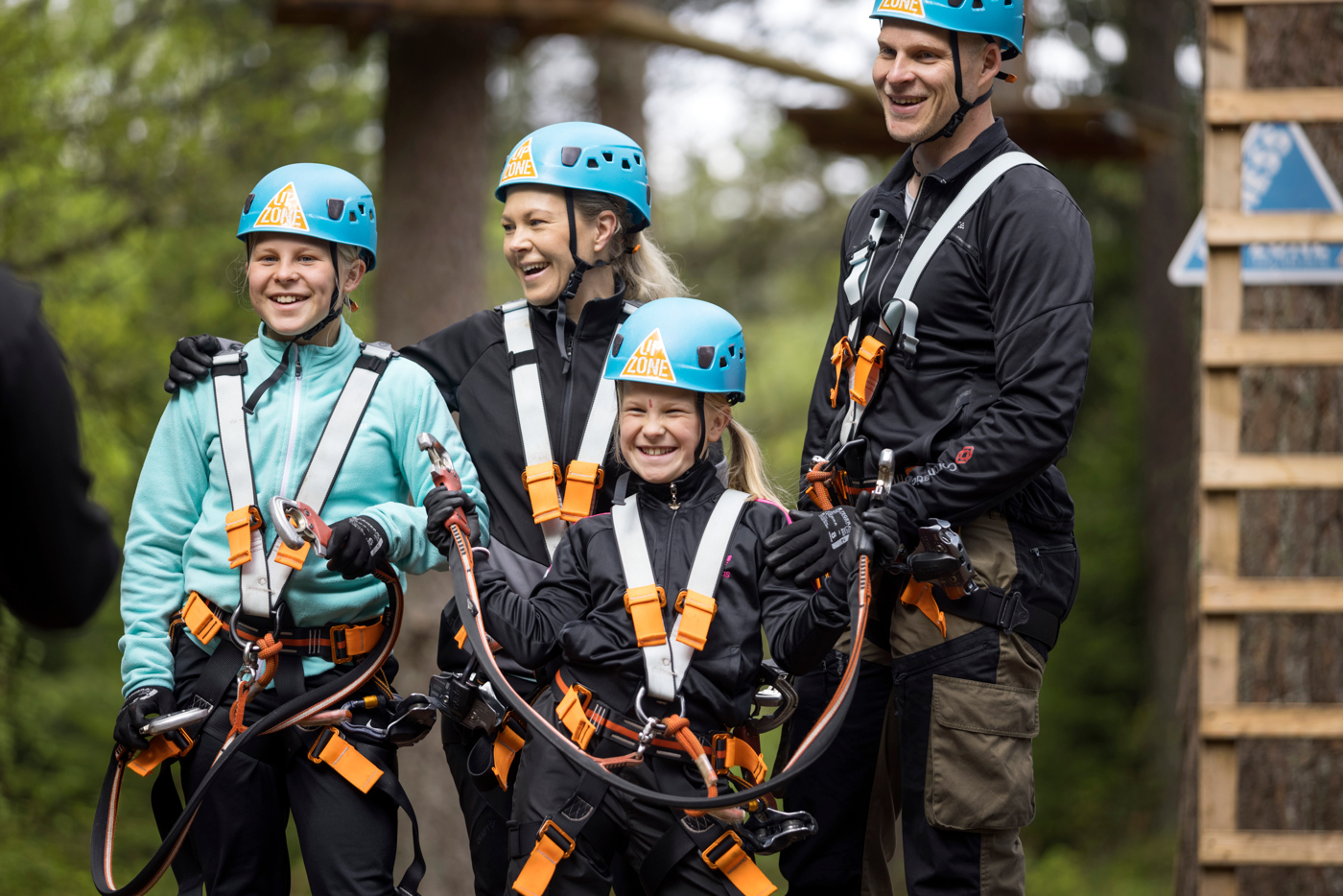 En skrattande familj står med klättrarutrustning redo att börja klättra upp i en höghöjdsbana. De har blå hjälmar och orange selar. 