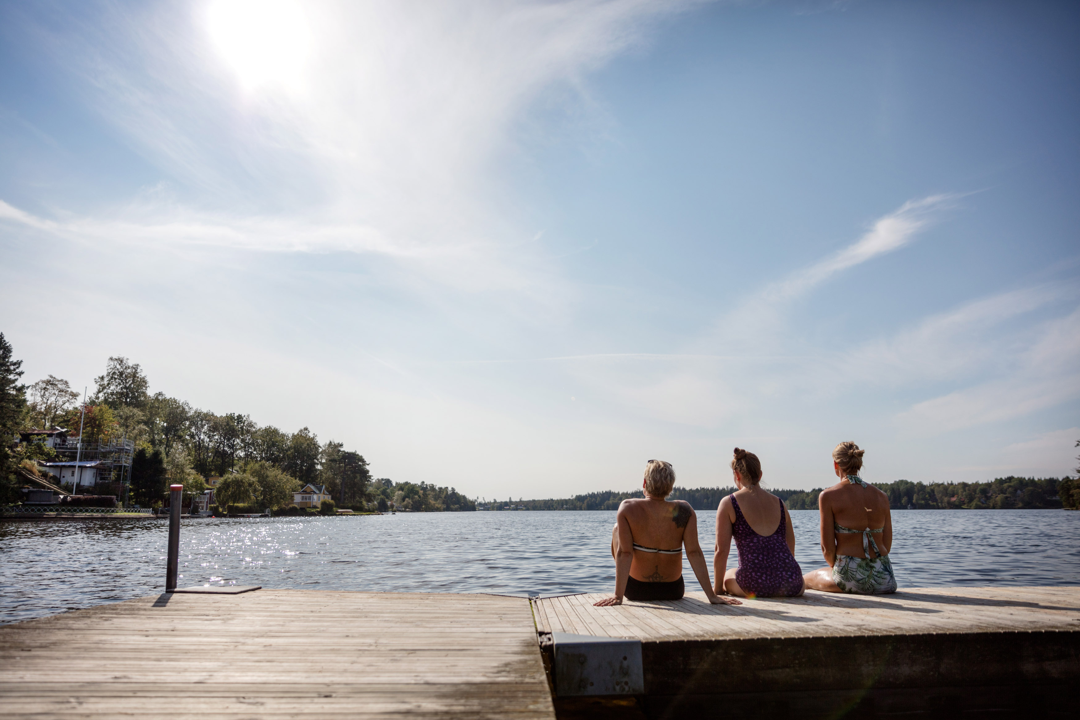 Tre kvinnor sitter på en brygga med ryggen mot kameran. Sjön i bakgrunden.