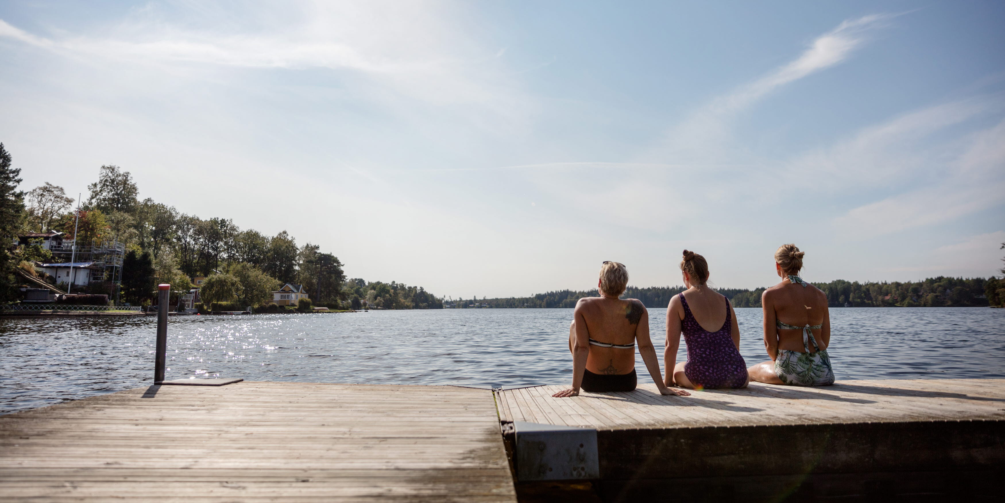 Tre kvinnor sitter på en brygga med ryggen mot kameran. Sjön i bakgrunden.