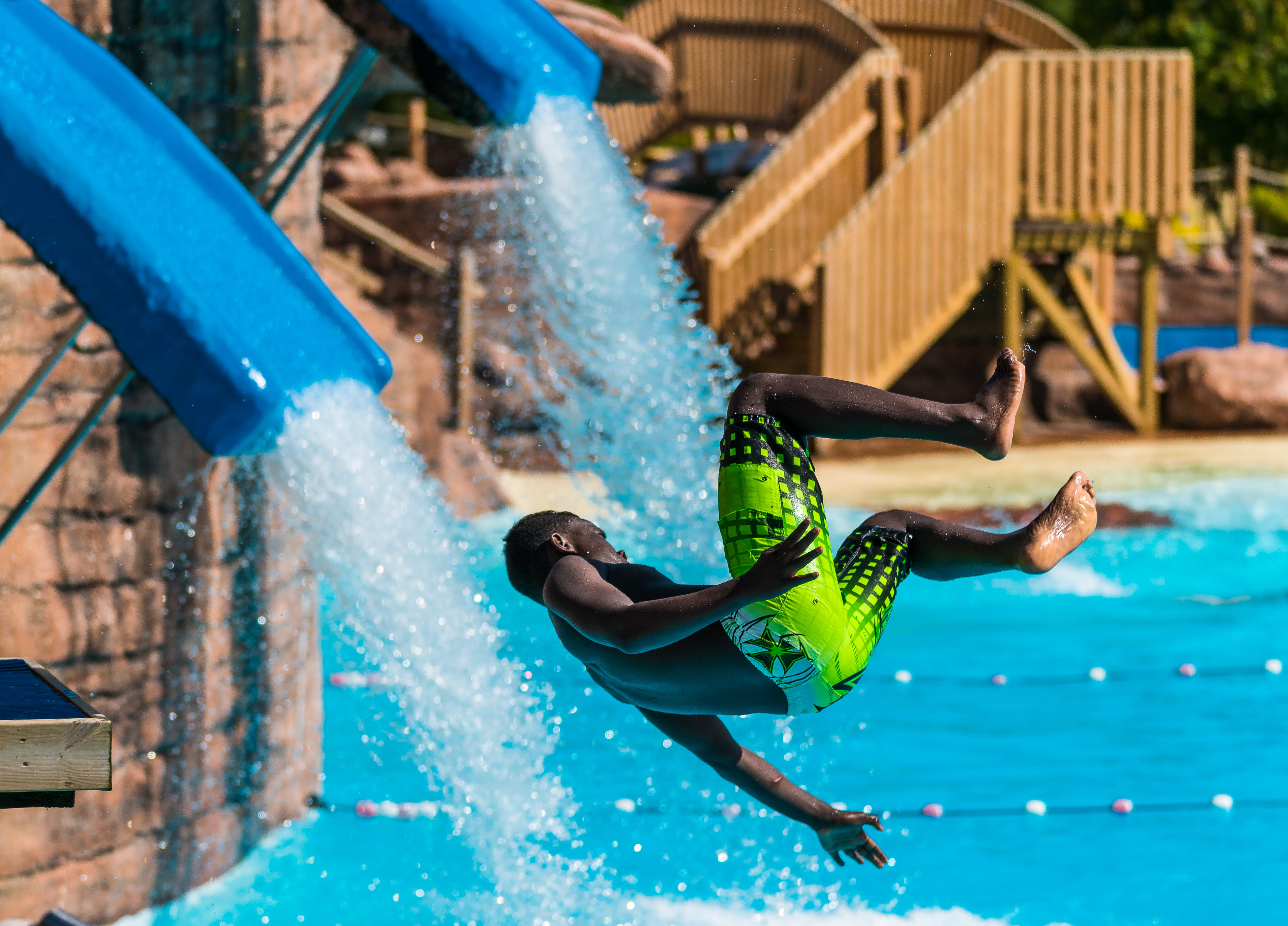 Ett barn flyger i luften ner från en vattenrutschkana. Snart landar han i en pool. Han har gröna badbyxor på sig.