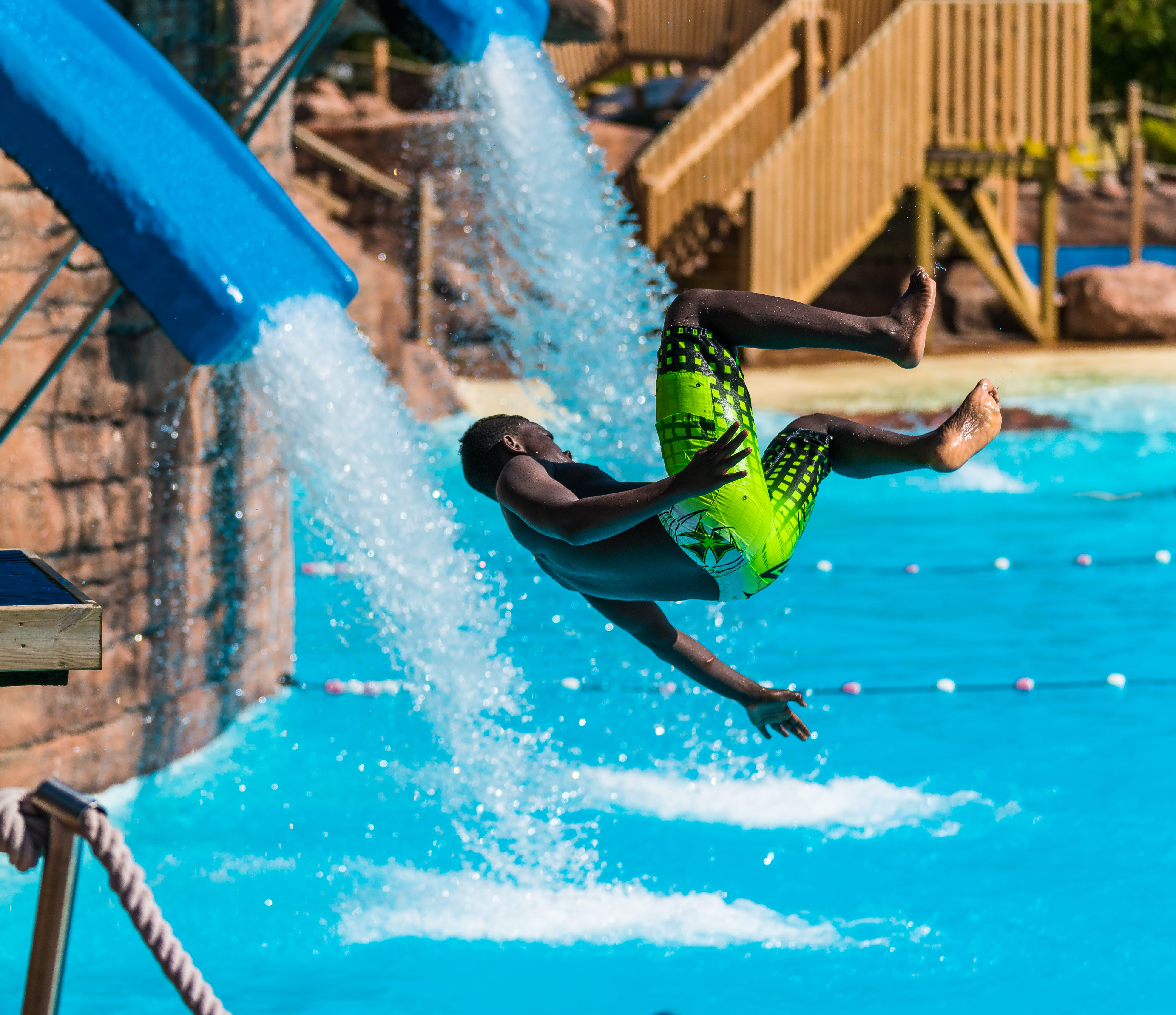 Ett barn flyger i luften ner från en vattenrutschkana. Snart landar han i en pool. Han har gröna badbyxor på sig.
