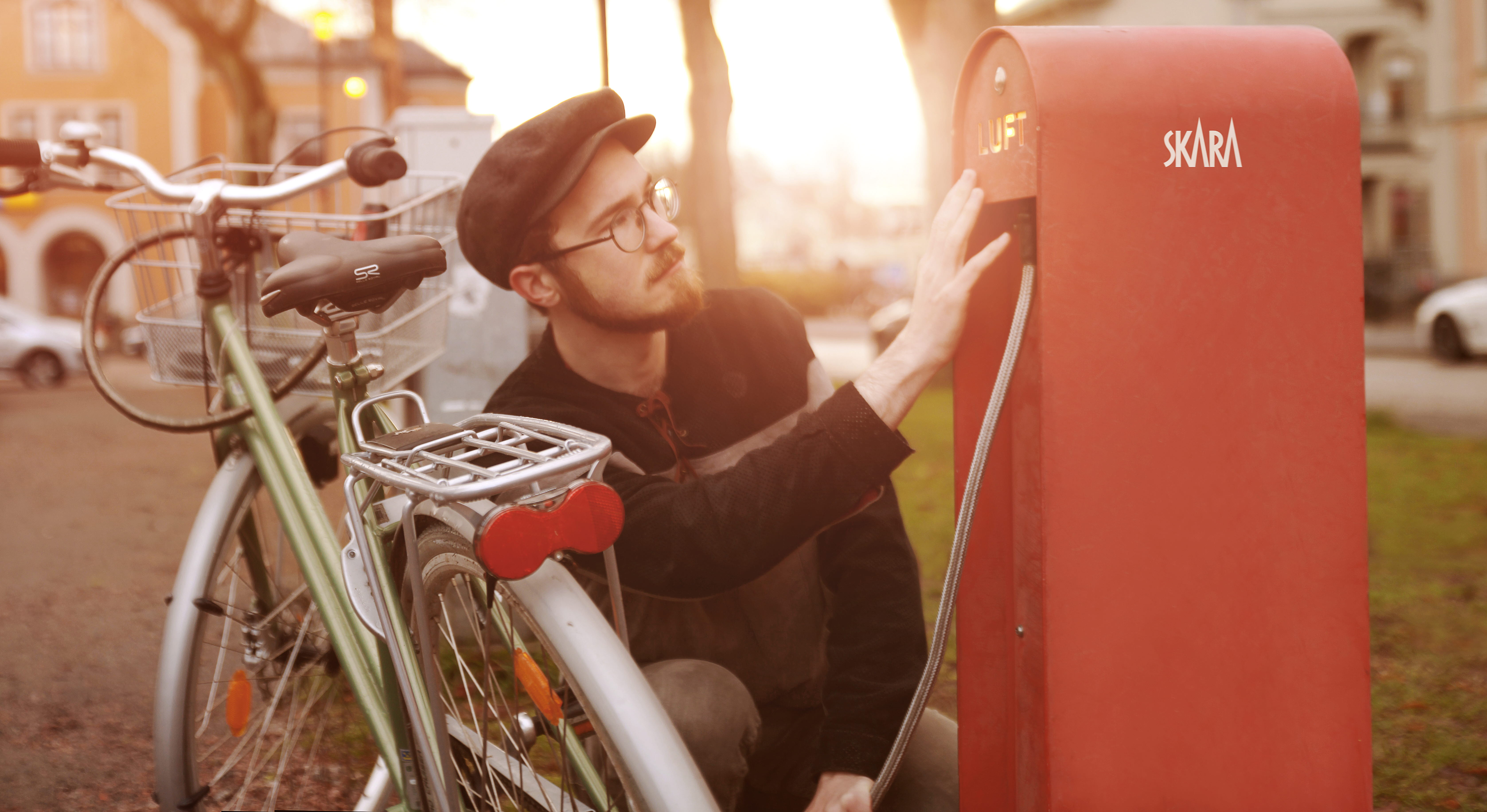 Man med glasögon och keps som pumpar sin cykel vid en stor röd cykelpump i plåt. 