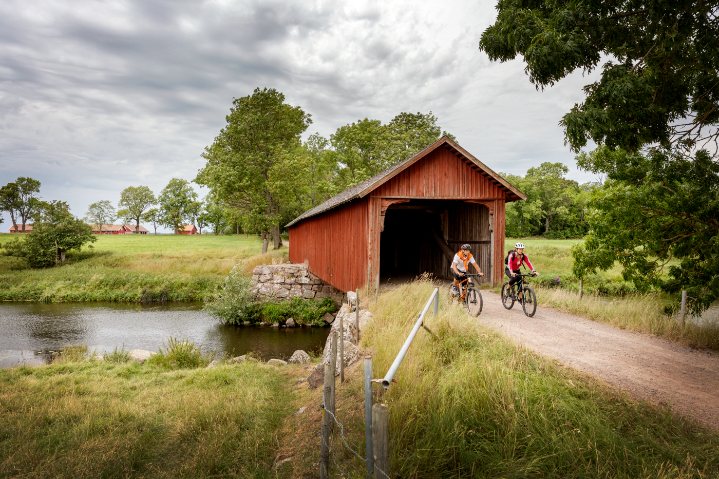 A woman and a man ride a mountain bike on a gravel road through a covered bridge next to a river.
