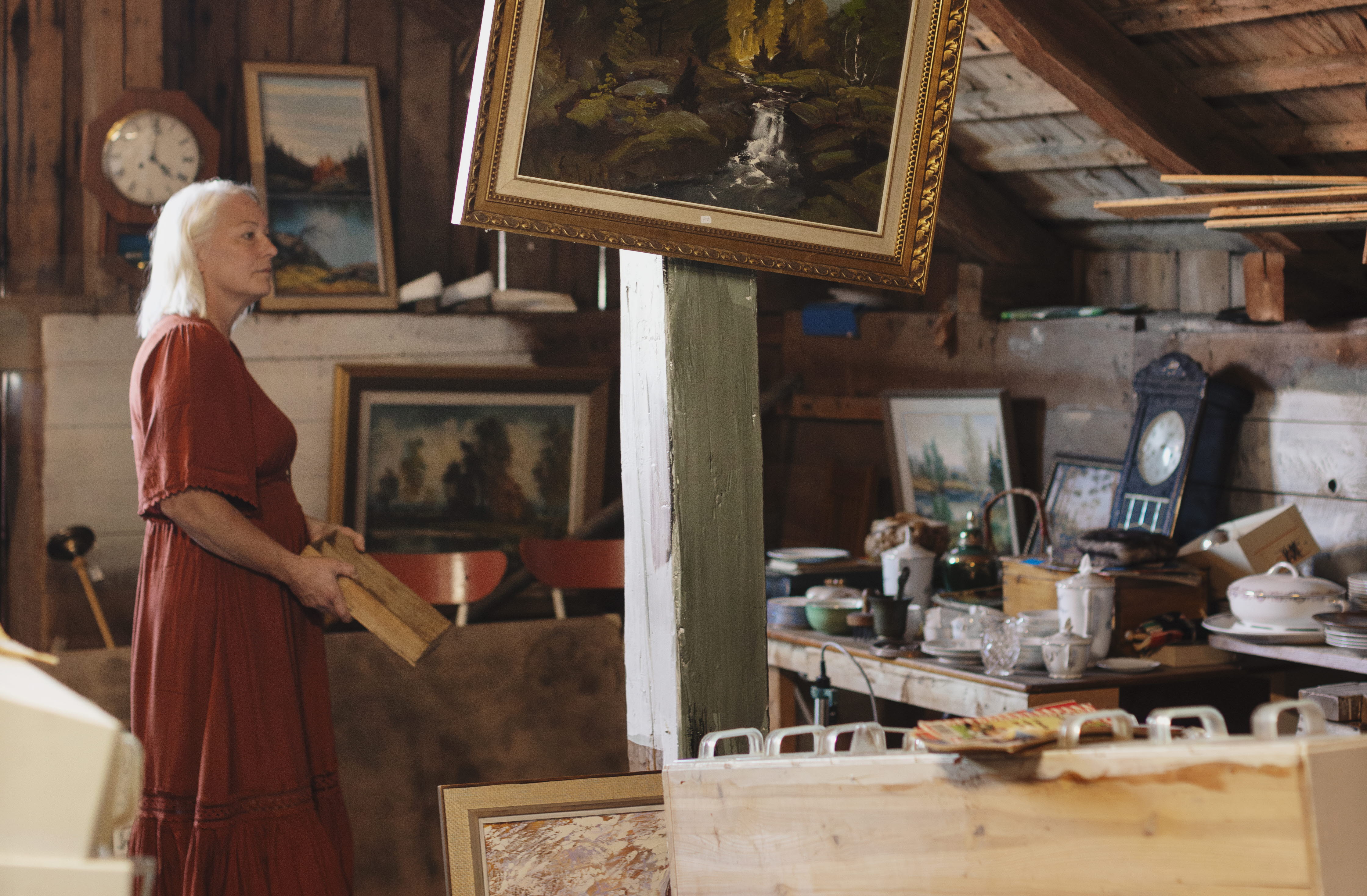 En kvinna i röd klänning är på en loppis i en lada. På en bärande stolpe hänger en sned tavla med guldram.