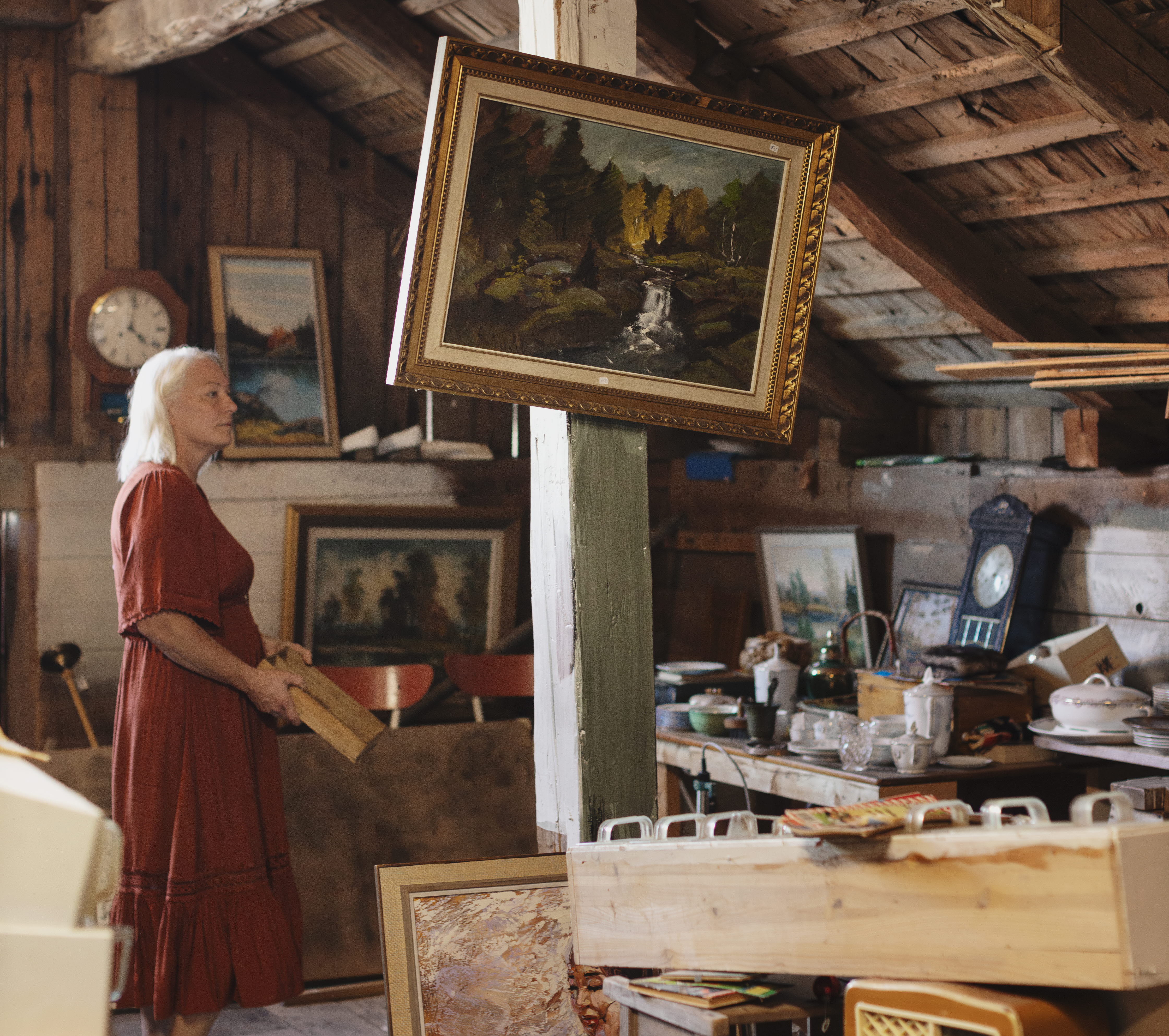 En kvinna i röd klänning är på en loppis i en lada. På en bärande stolpe hänger en sned tavla med guldram.