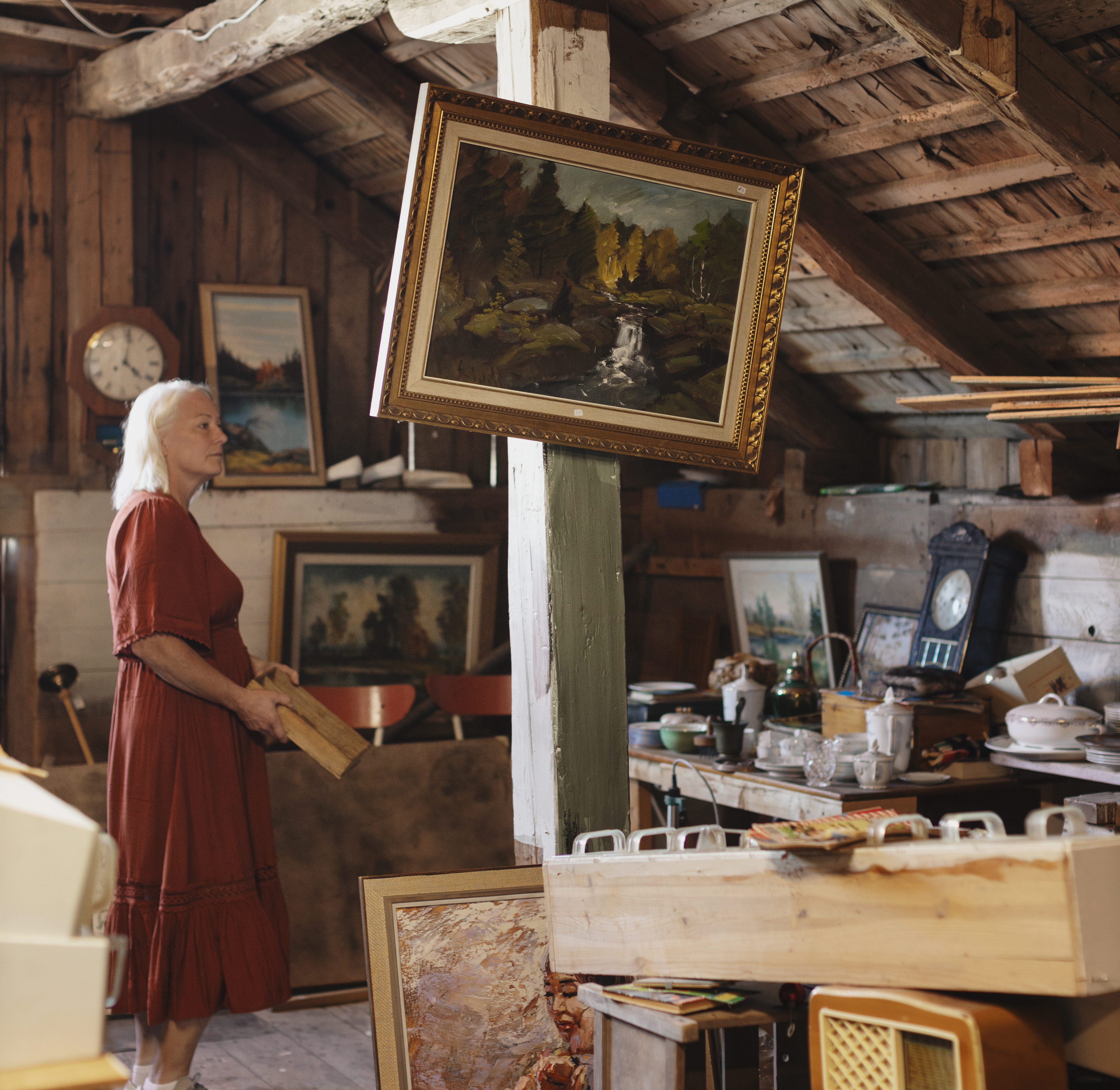 En kvinna i röd klänning är på en loppis i en lada. På en bärande stolpe hänger en sned tavla med guldram.