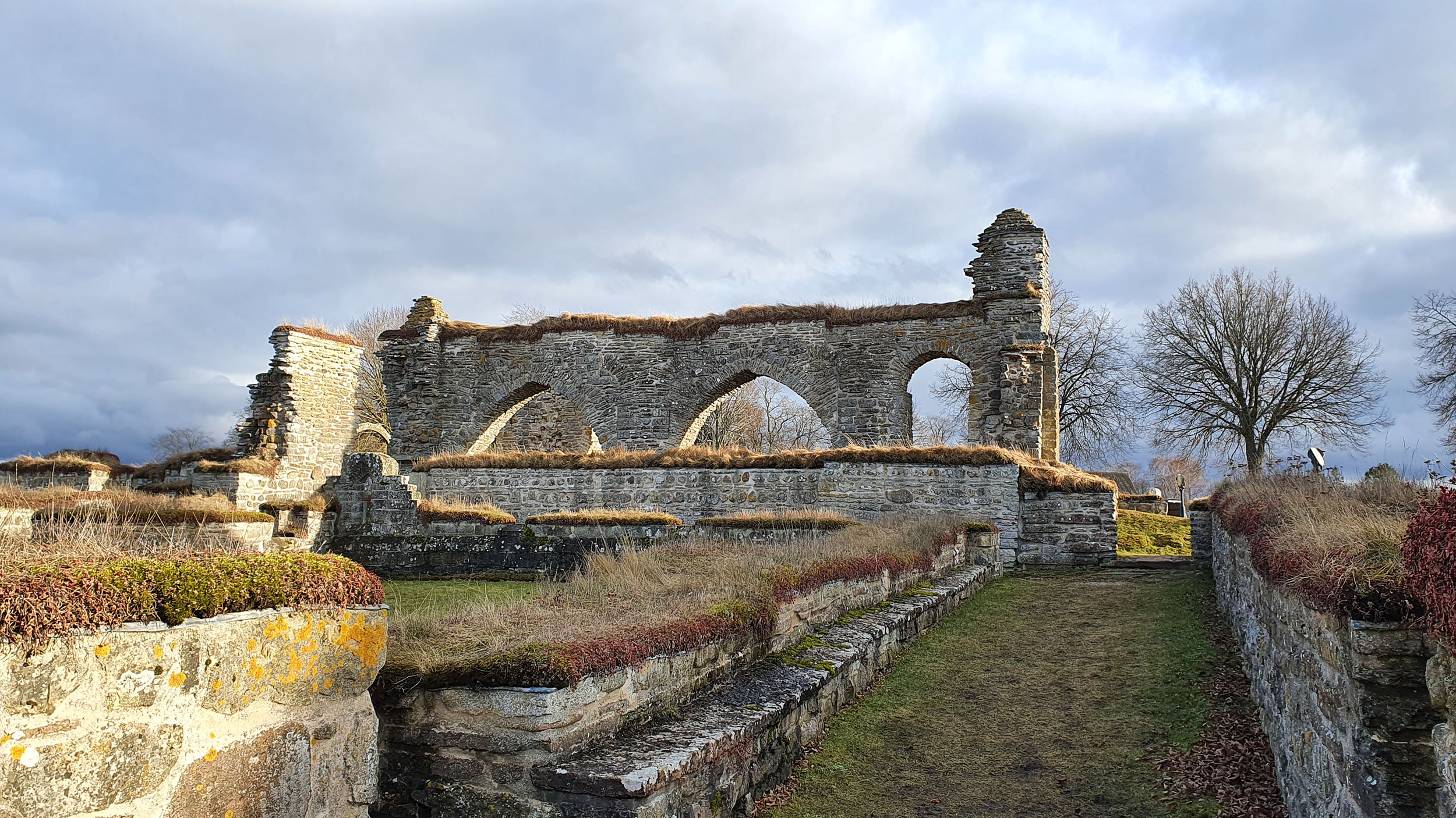 Ett landskap med en klosterruin med låga murar och en vägg med tre valv. På himlen syns moln och i bakgrunden ett träd utan löv. 
