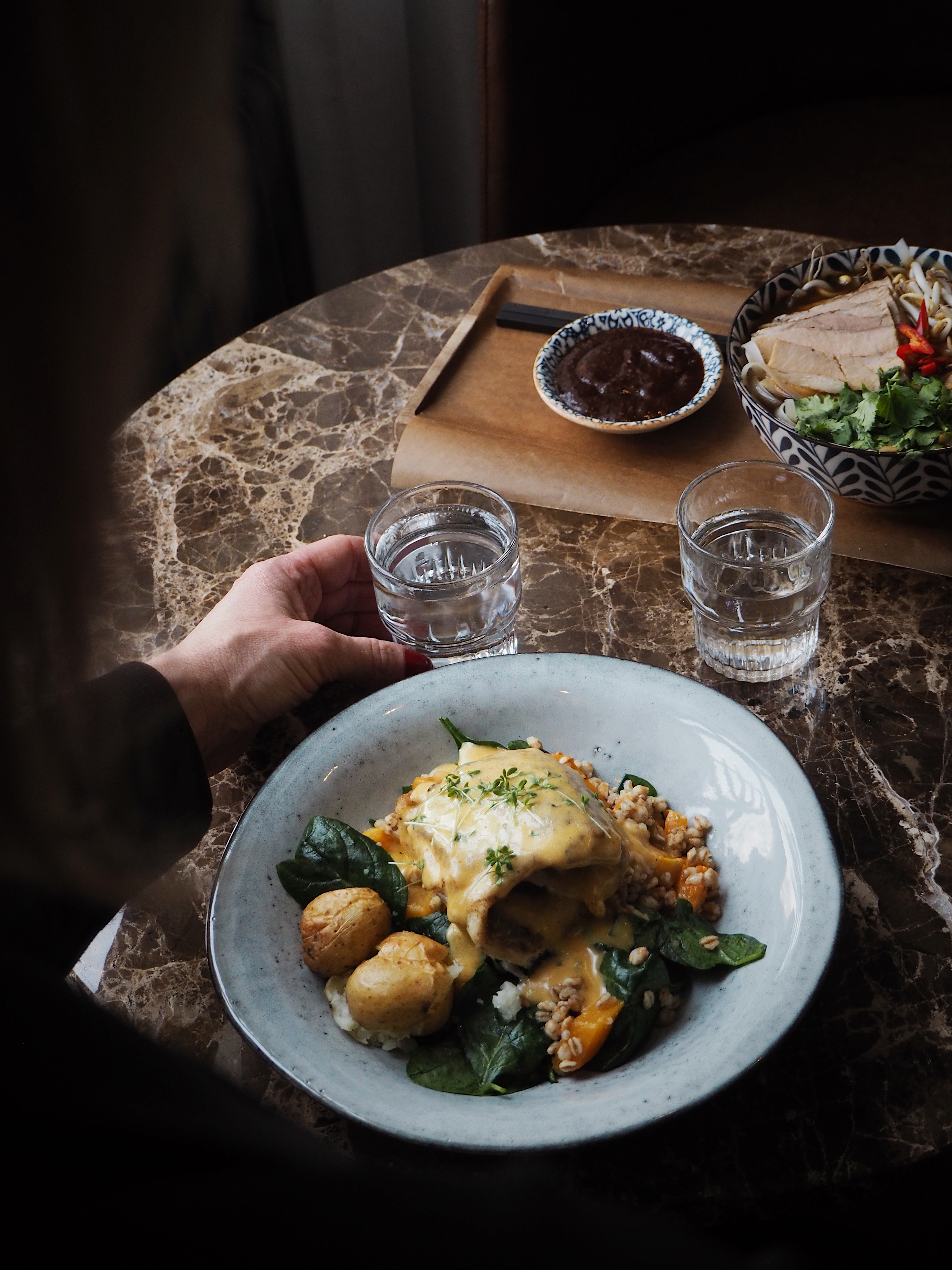 Någon sitter och äter vid ett runt bord och håller i ett dricksglas. En tallrik med mat och tillbehör vid sidan om.