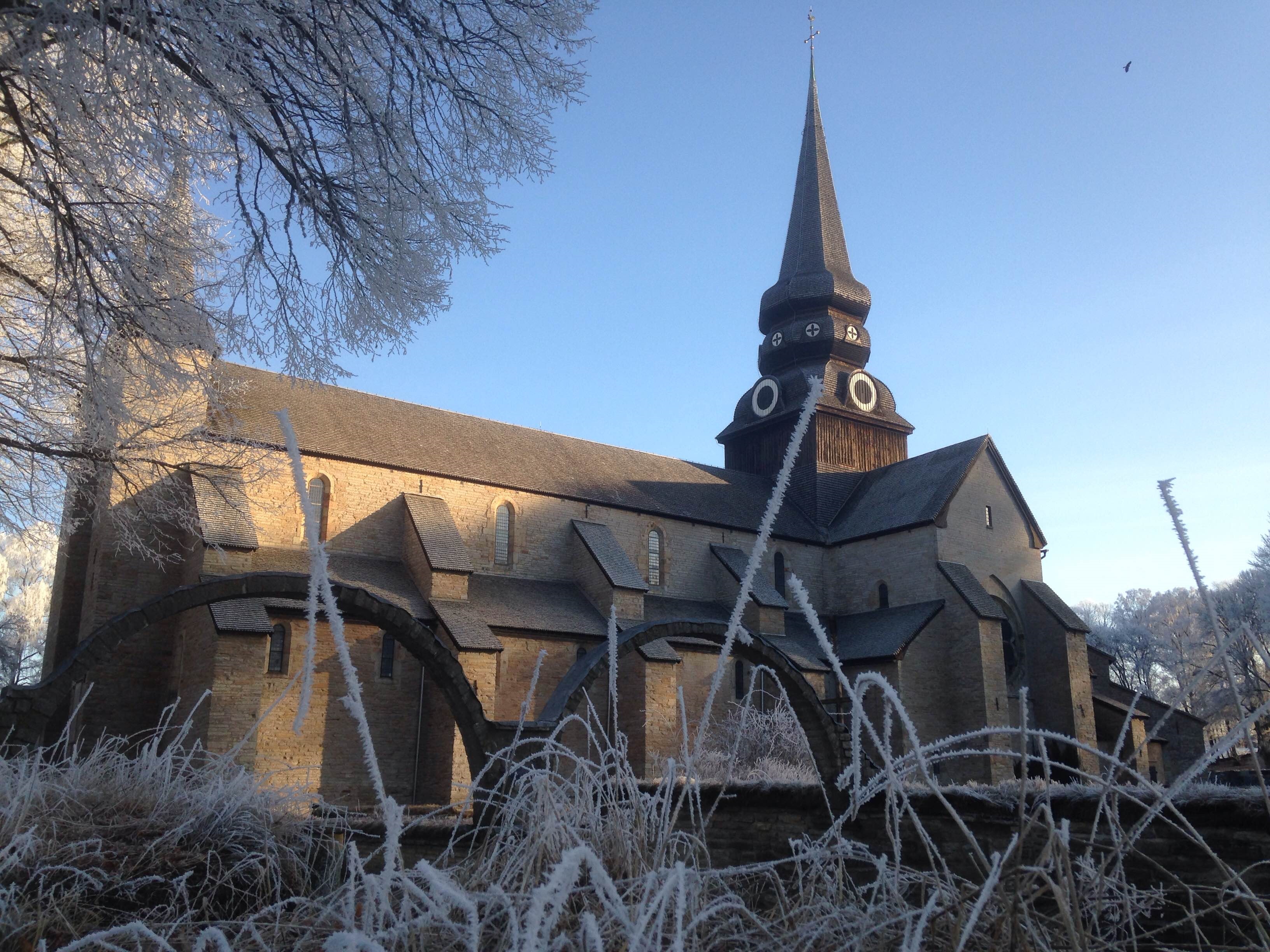 En stor grå stenkyrka med ett tjärat torn. I förgrunden syns två stenbågar från ett gammalt kloster och frostiga grässtrån.