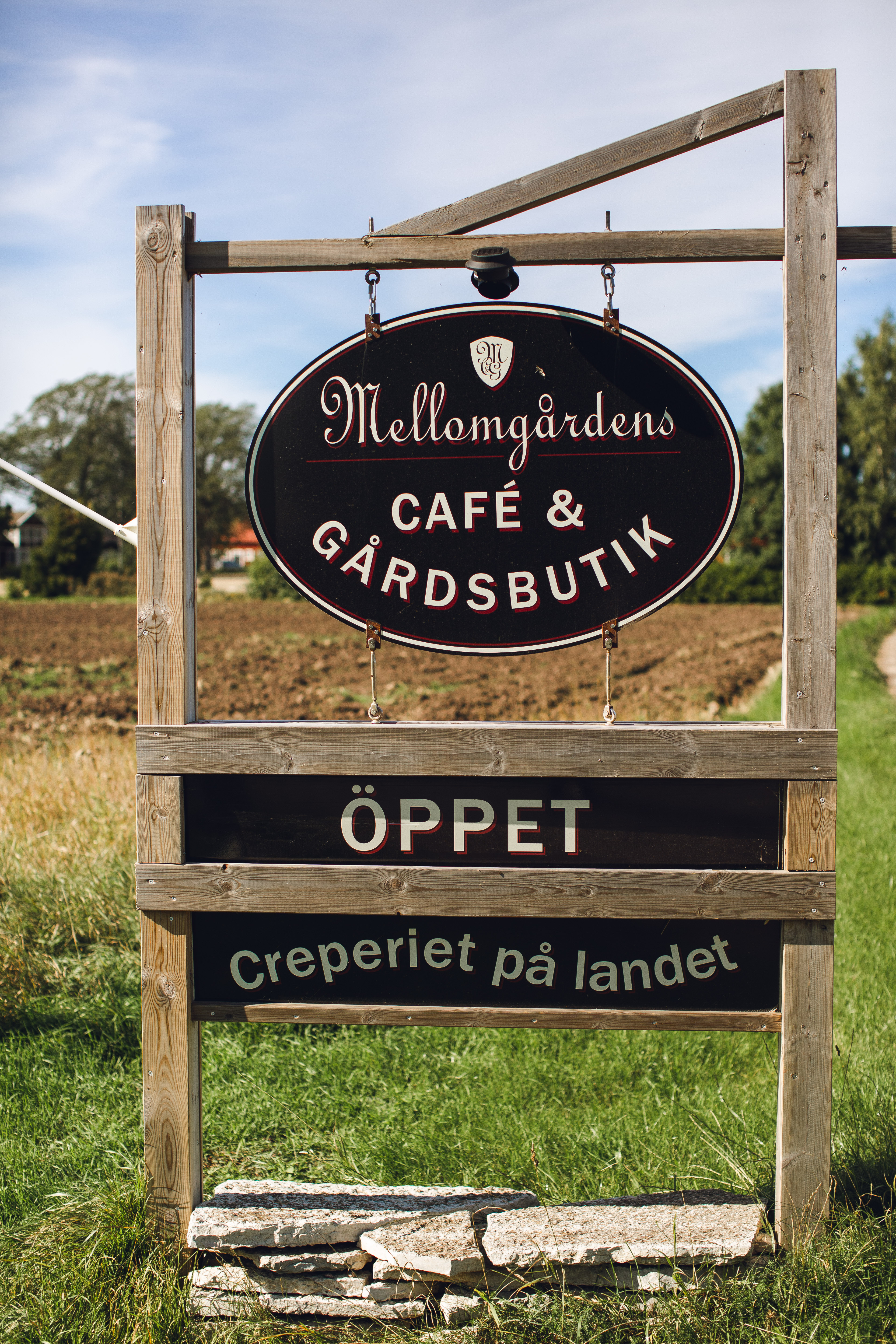 En träskylt där det står Mellomgårdens Café & Gårdsbutik, Öppet, Creperiet på landet.