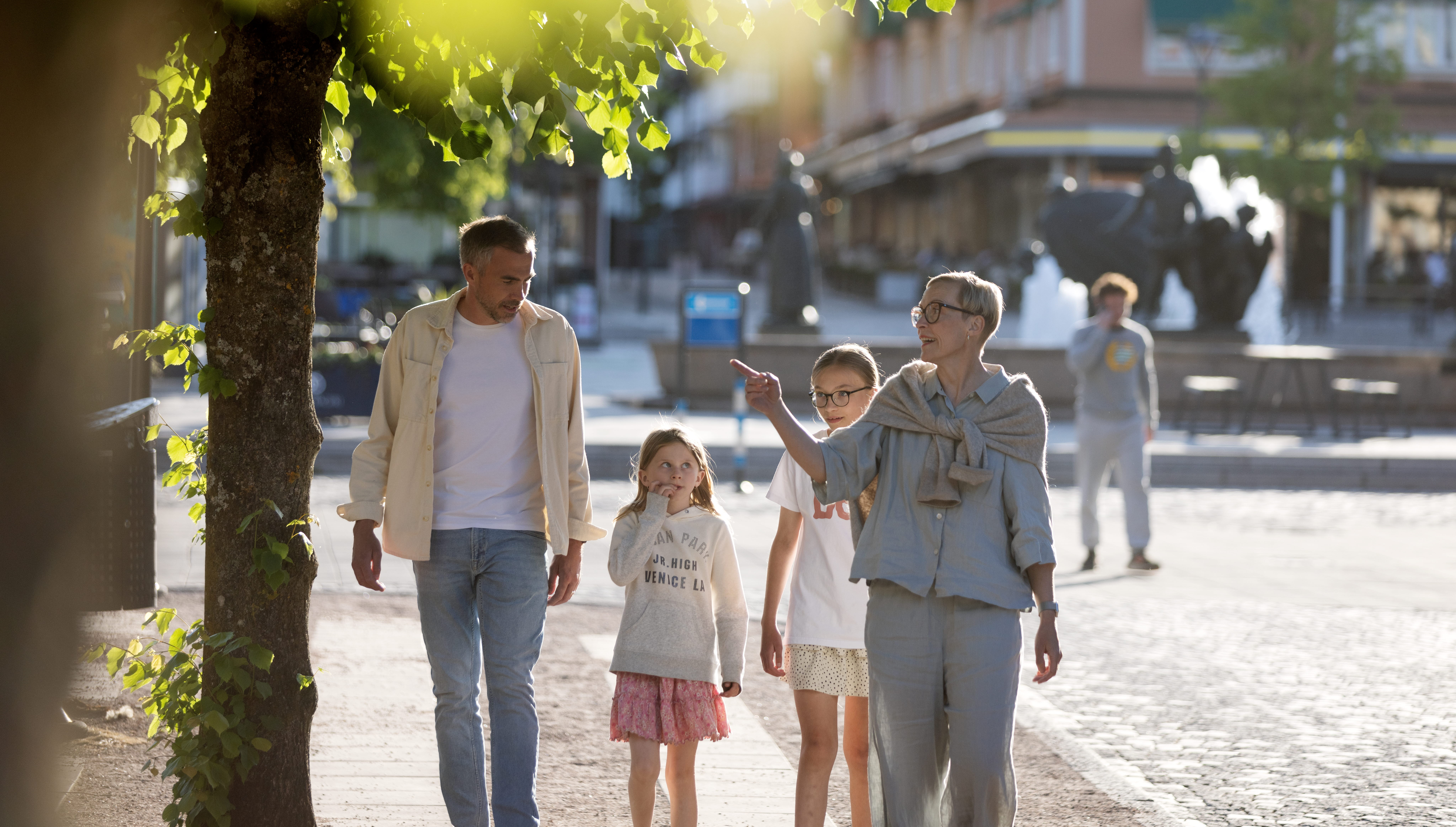 En man, en flicka, en ung tjej och en kvinna promenerar i en stad med grönskande träd framför och byggnader bakom sig.