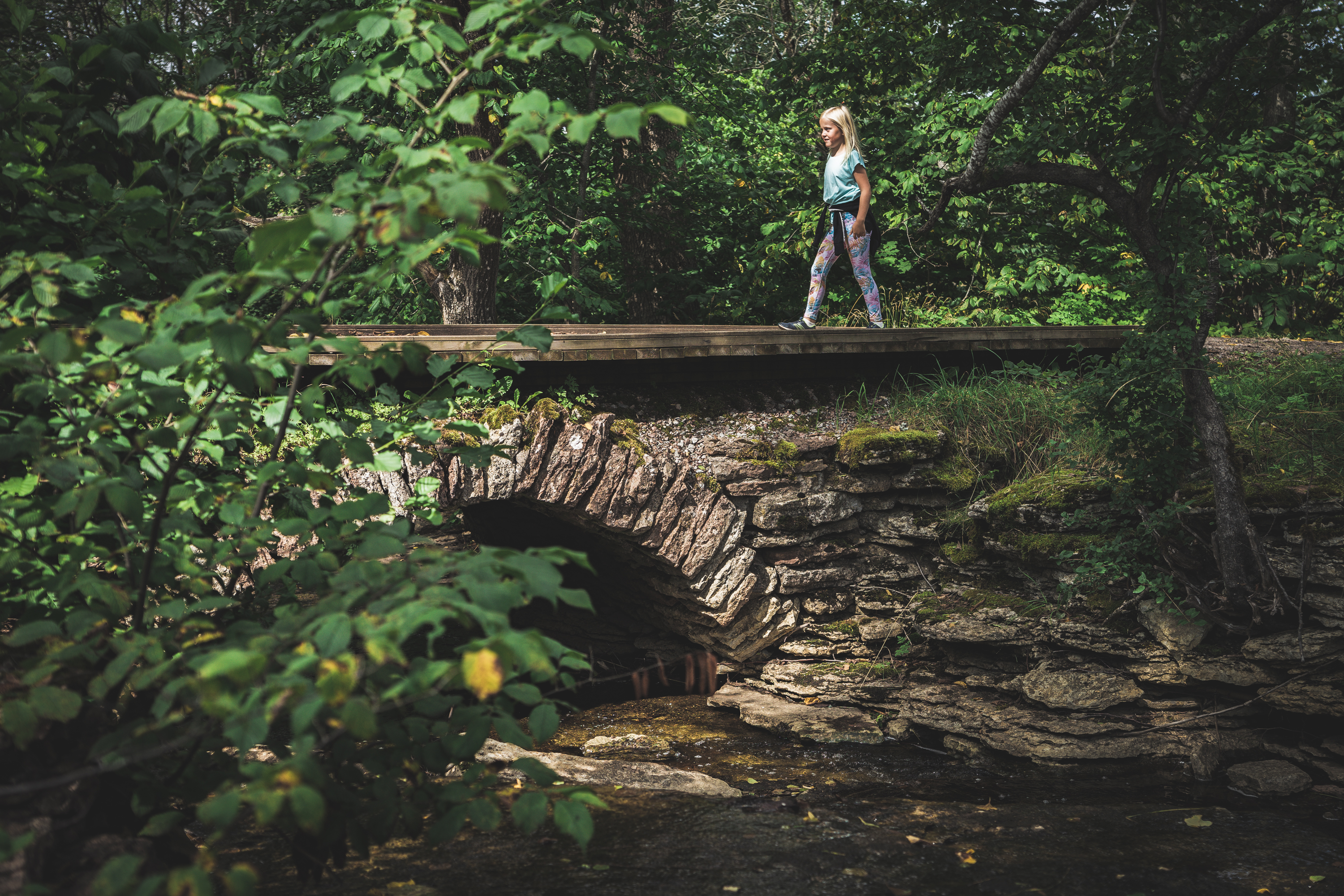A child dressed in light clothes is walking over an arched stone bridge. The trees around the bridge are green.