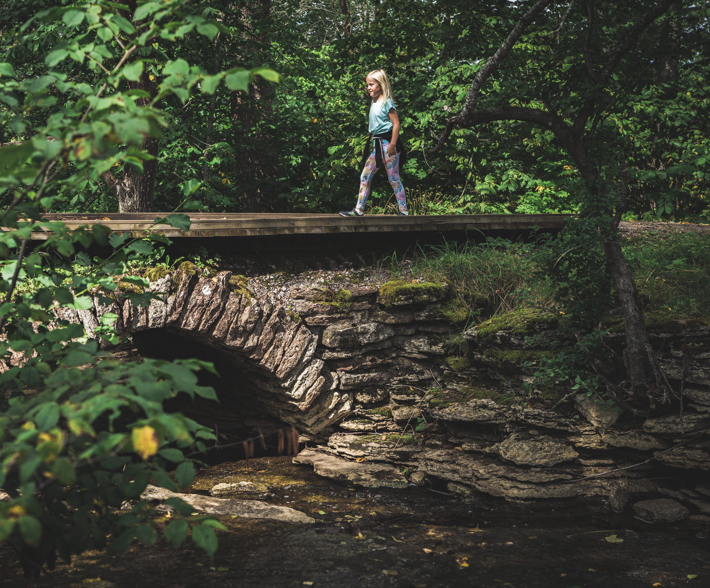 Ett barn i ljusa kläder går över en välvd stenbro. Träden runt bron grönskar. 