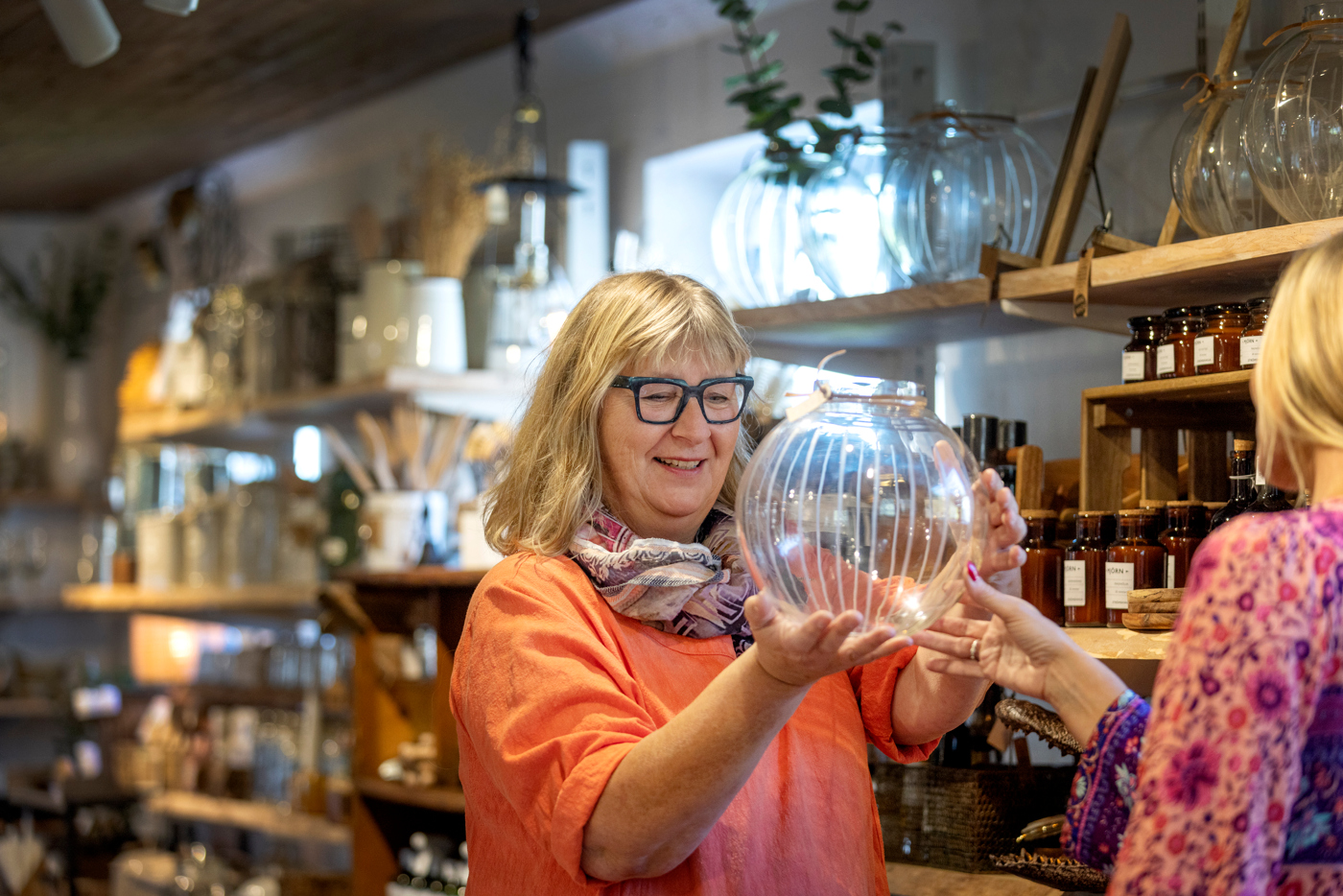 A woman in black glasses and a coral colored sweater looking at a glass vase in a shop.