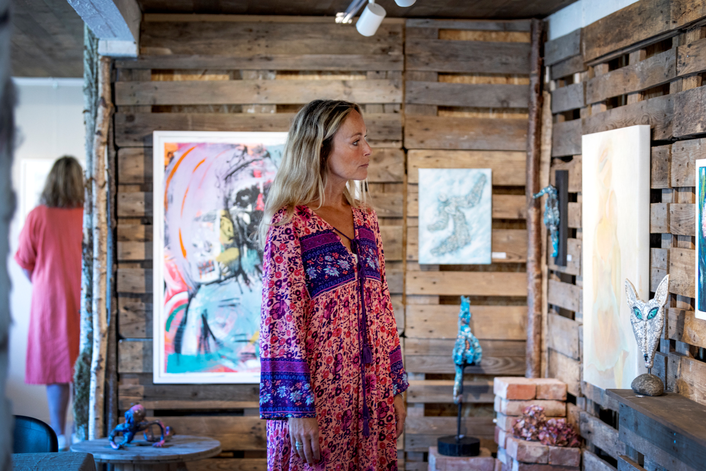 En kvinna i blont hår och mönstrad långklänning står och beundrar konst i en konsthall. 