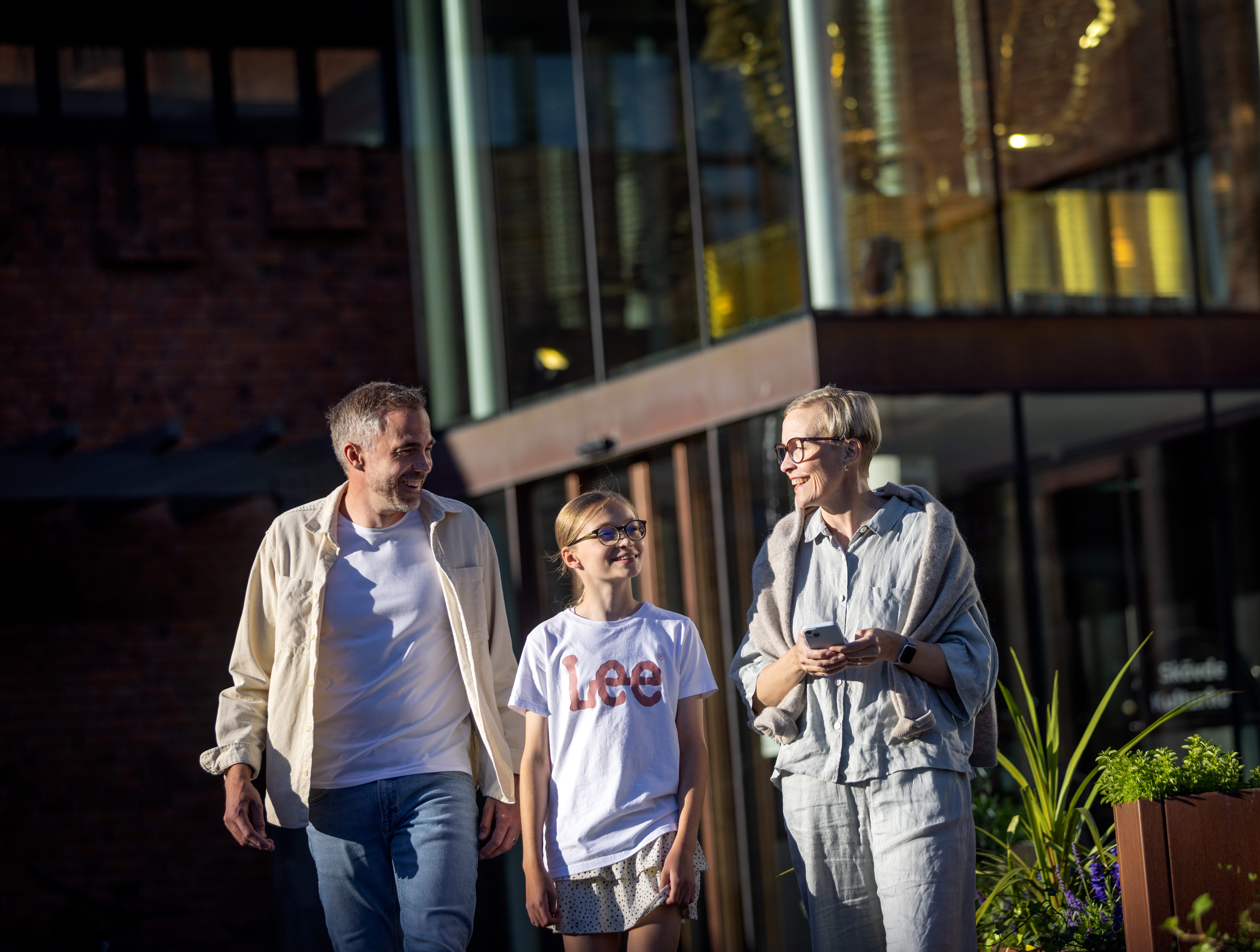 En man, en ung tjej och en kvinna ler och pratar med varandra utanför en byggnad.