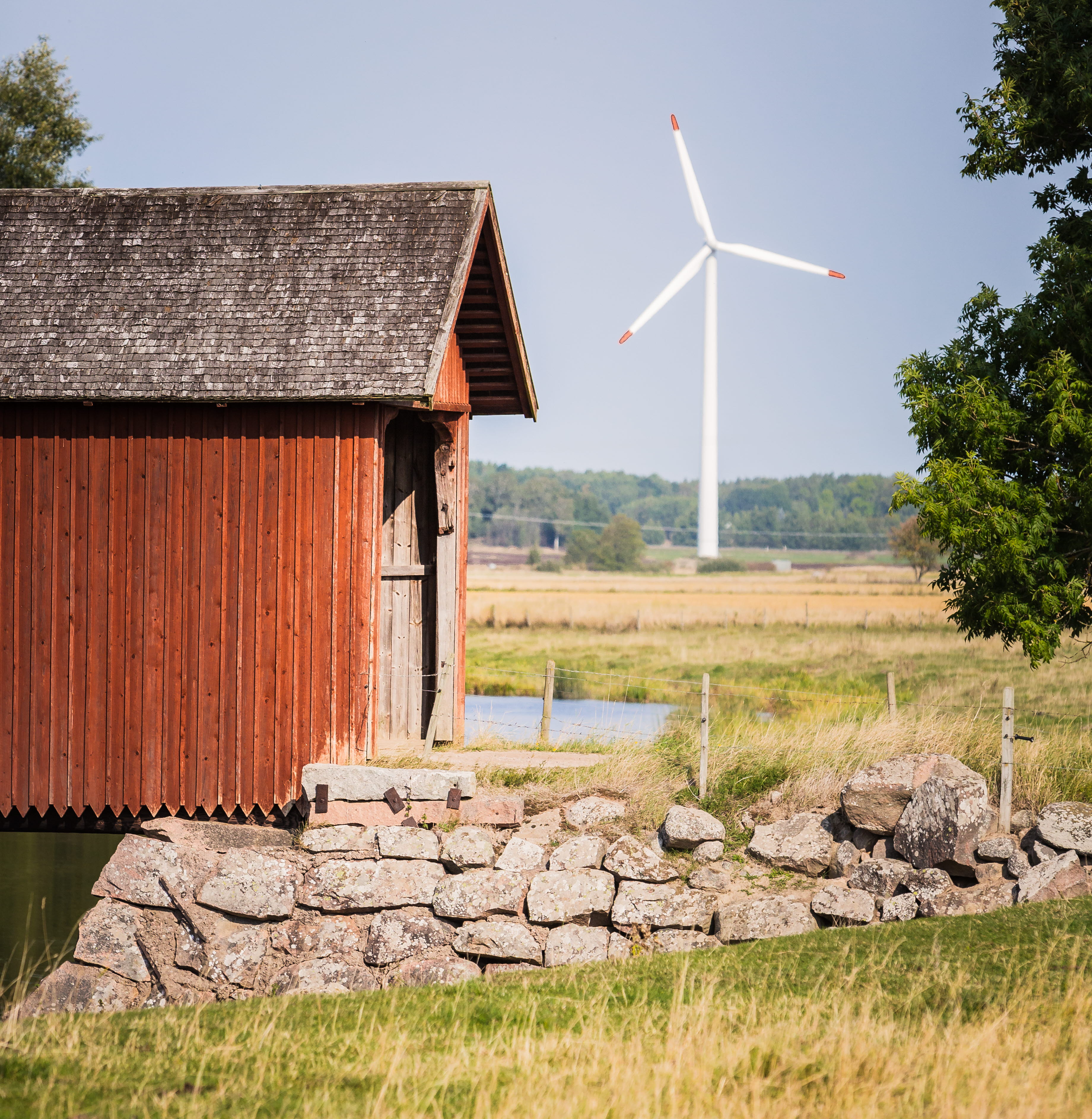 Rött hus över en å. Stenmur vid kanten och ett vindkraftverk i bakgrunden.