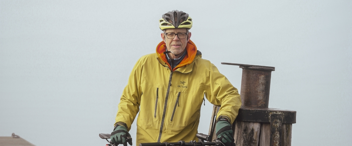 En man som har cykelhjälm på sig tittar in i kameran. Det är dimmigt bakom honom. Han har jacka på sig. 