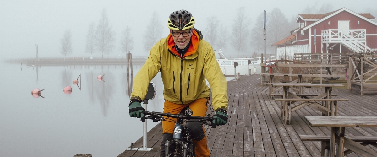 En man som har cykelhjälm på sig tittar in i kameran när han sitter på sin cykel. Det är dimmigt bakom honom. Han har jacka och glasögon på sig. 