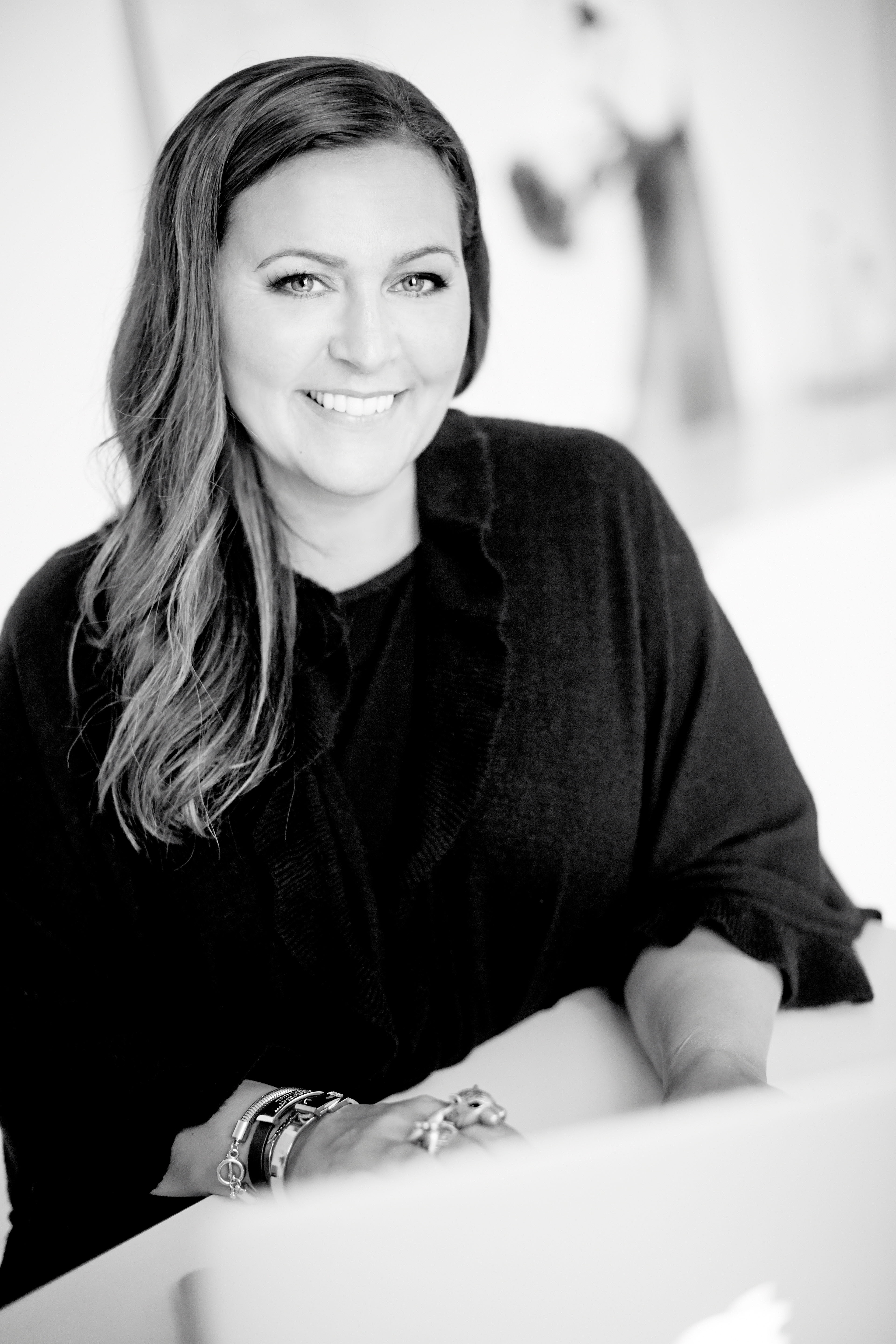 Svartvitt foto på leende person med långt brunt hår sittande vid skrivbord.