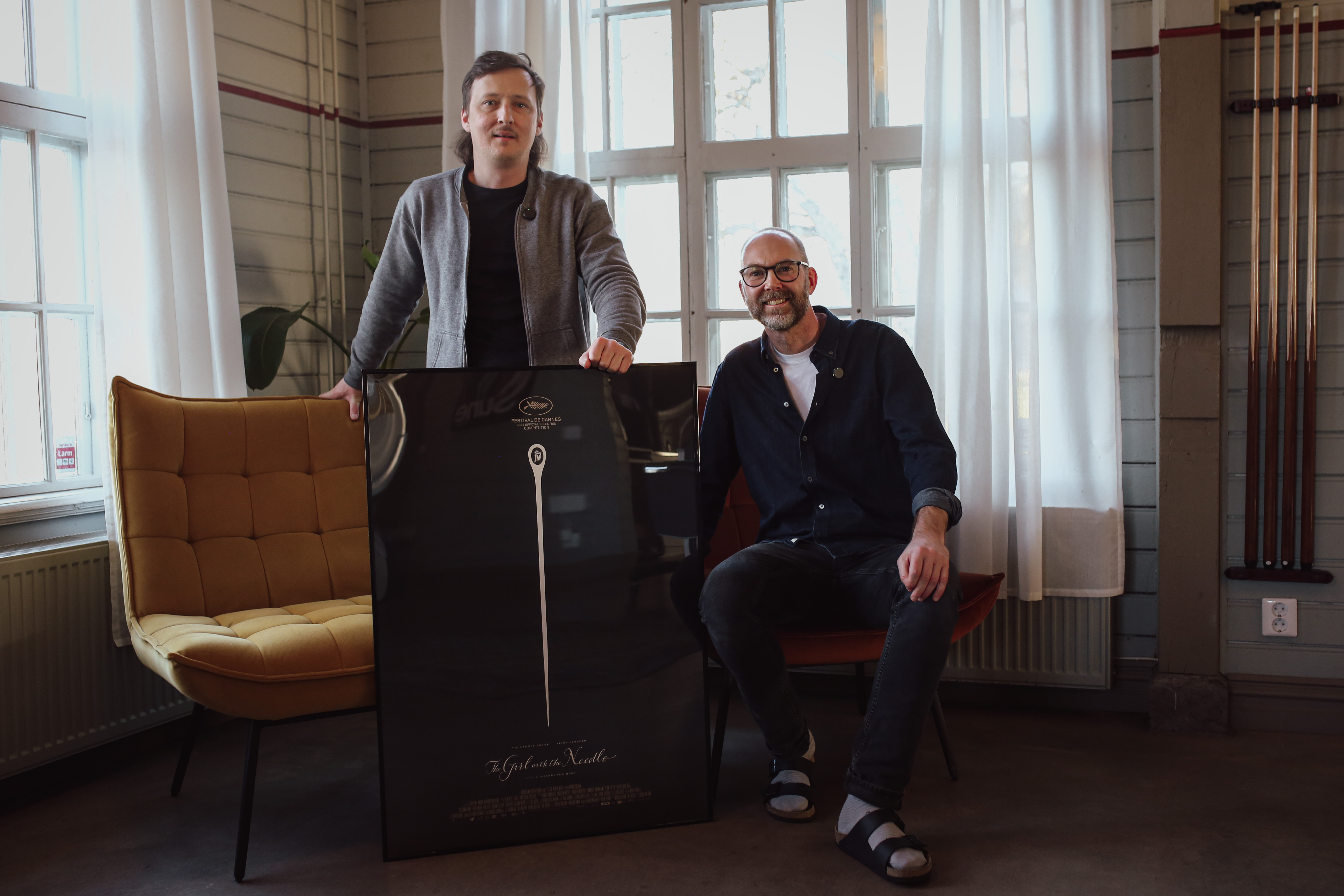 Två personer, den som är till vänster står upp och den som är till höger sitter ner i en fåtölj. Personen som står upp håller en tavla som är placerad framför på golvet.