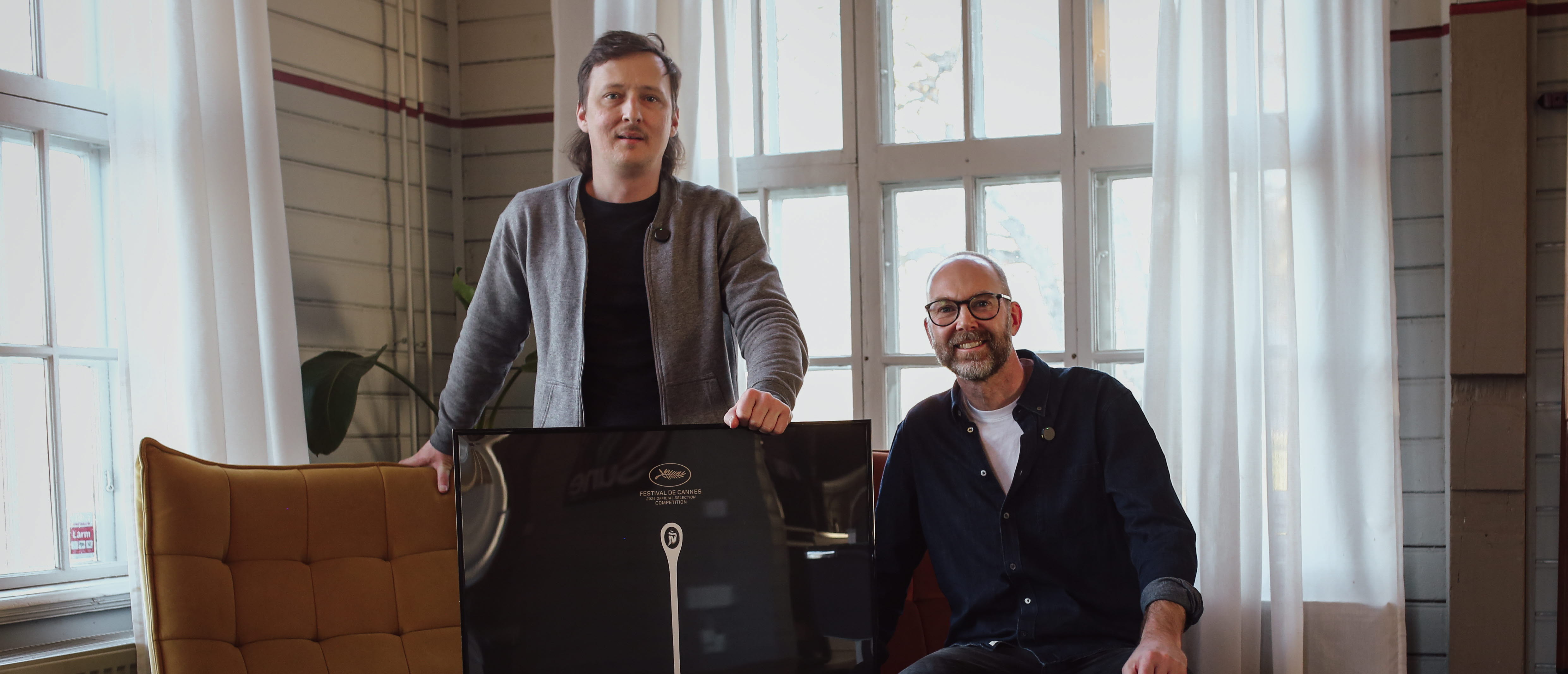 Två personer, den som är till vänster står upp och den som är till höger sitter ner i en fåtölj. Personen som står upp håller en tavla som är placerad framför på golvet.