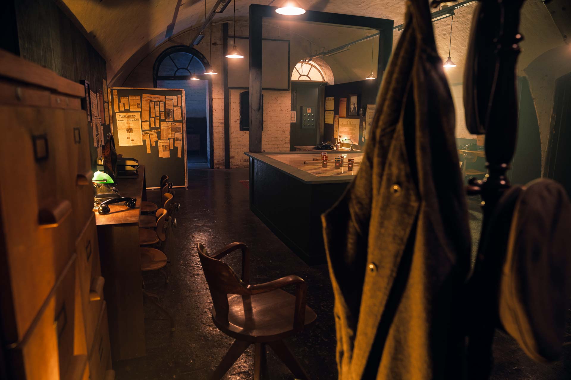 Ett mörkt kontorsrum från 1900-talets början med några få ljuskällor som lyser upp rummet. I förgrunden syns en herrbetjänt med en militäruniform. En stol av trä står på golvet och bakom den syns skrivbord och ett stort bord med en kartbild som toppskiva. 