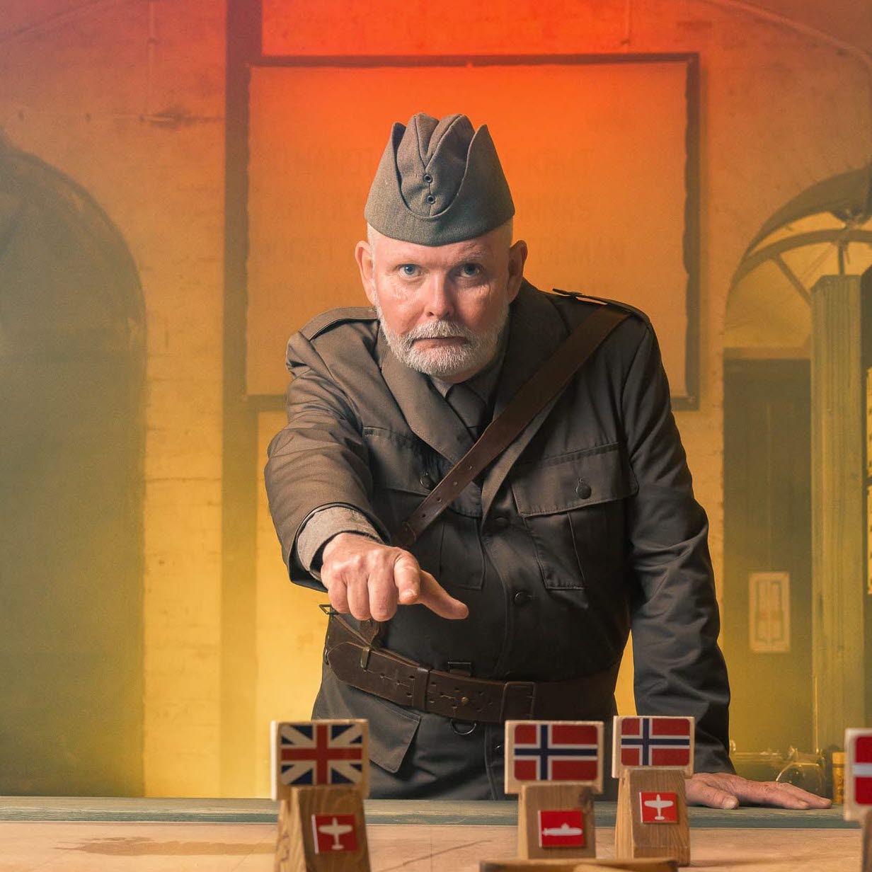 En man i uniform står och pekar mot kameran och ett bord med en karta som toppskiva. På skivan står flaggmarkörer utspridda med Storbritanniens och Norges flaggor. 