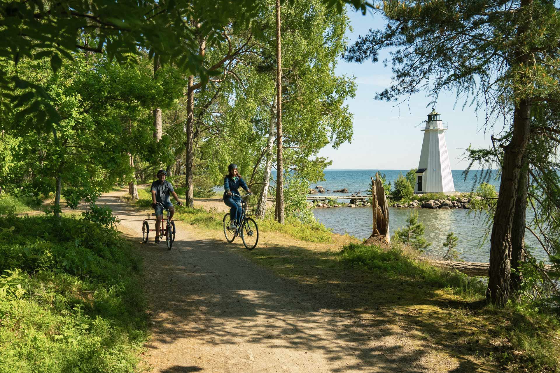Två personer cyklar på en stig bredvid vatten där en ser en fyr i bakgrunden till höger. Den ena personen cyklar på en vuxentrehjuling.