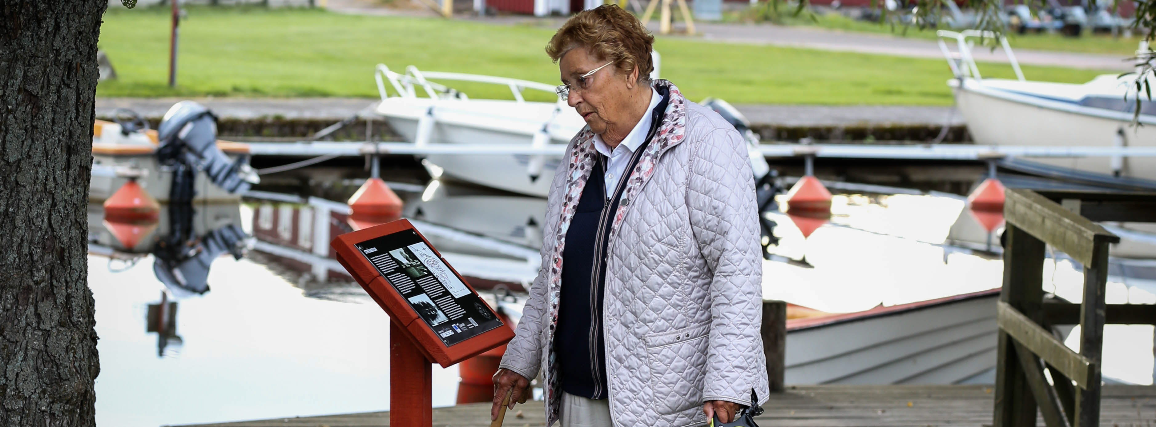 En äldre person med käpp och hund kopplad intill sig står och läser på en informationsskylt. Skylten är placerad intill ett träd och i bakgrunden syns en smal kanal där båtar ligger förankrade.