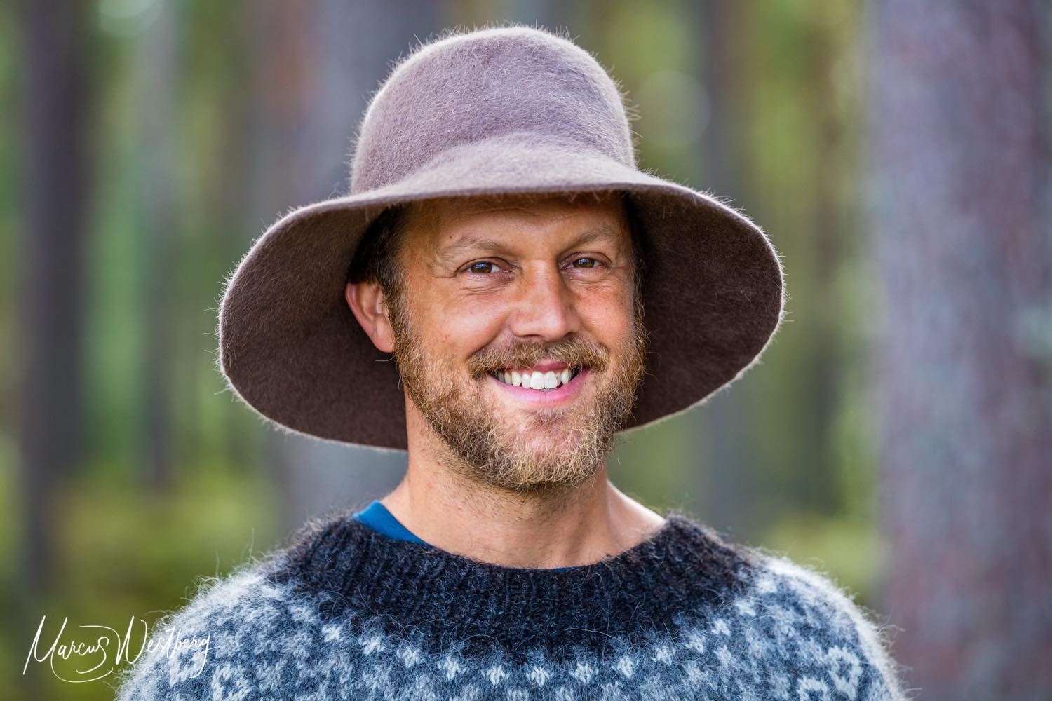 Leende person med stickad tröja och hatt tittar in i kameran. Personen befinner sig i en skog.