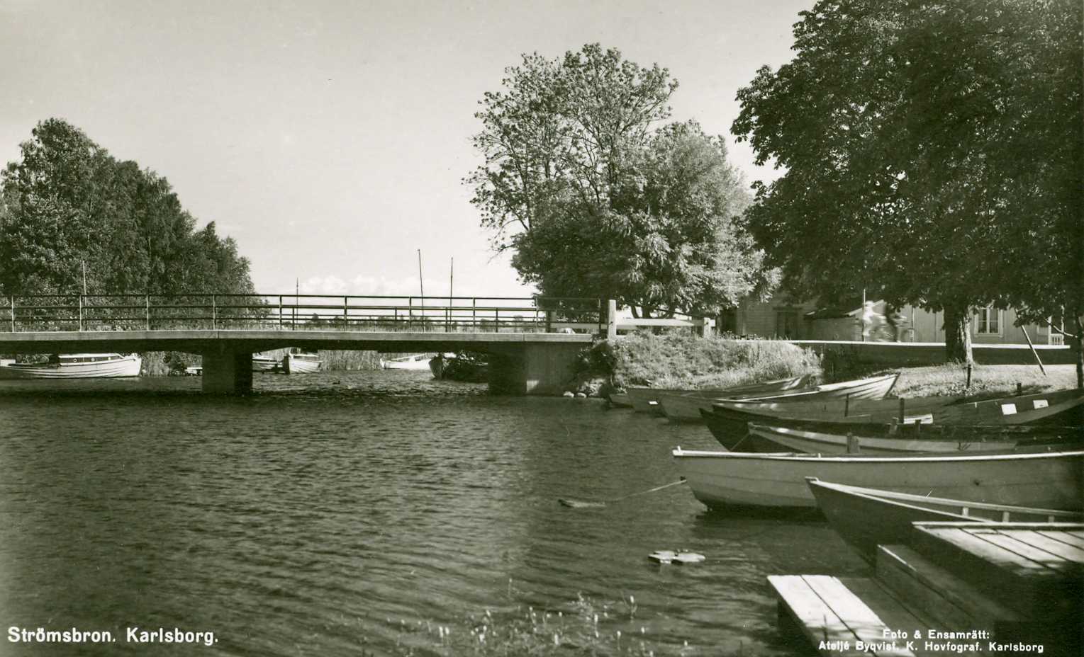 Black and white archive image of a canal with boats moored along the edge. Further in the distance, there is a bridge, Strömbron in Karlsborg.