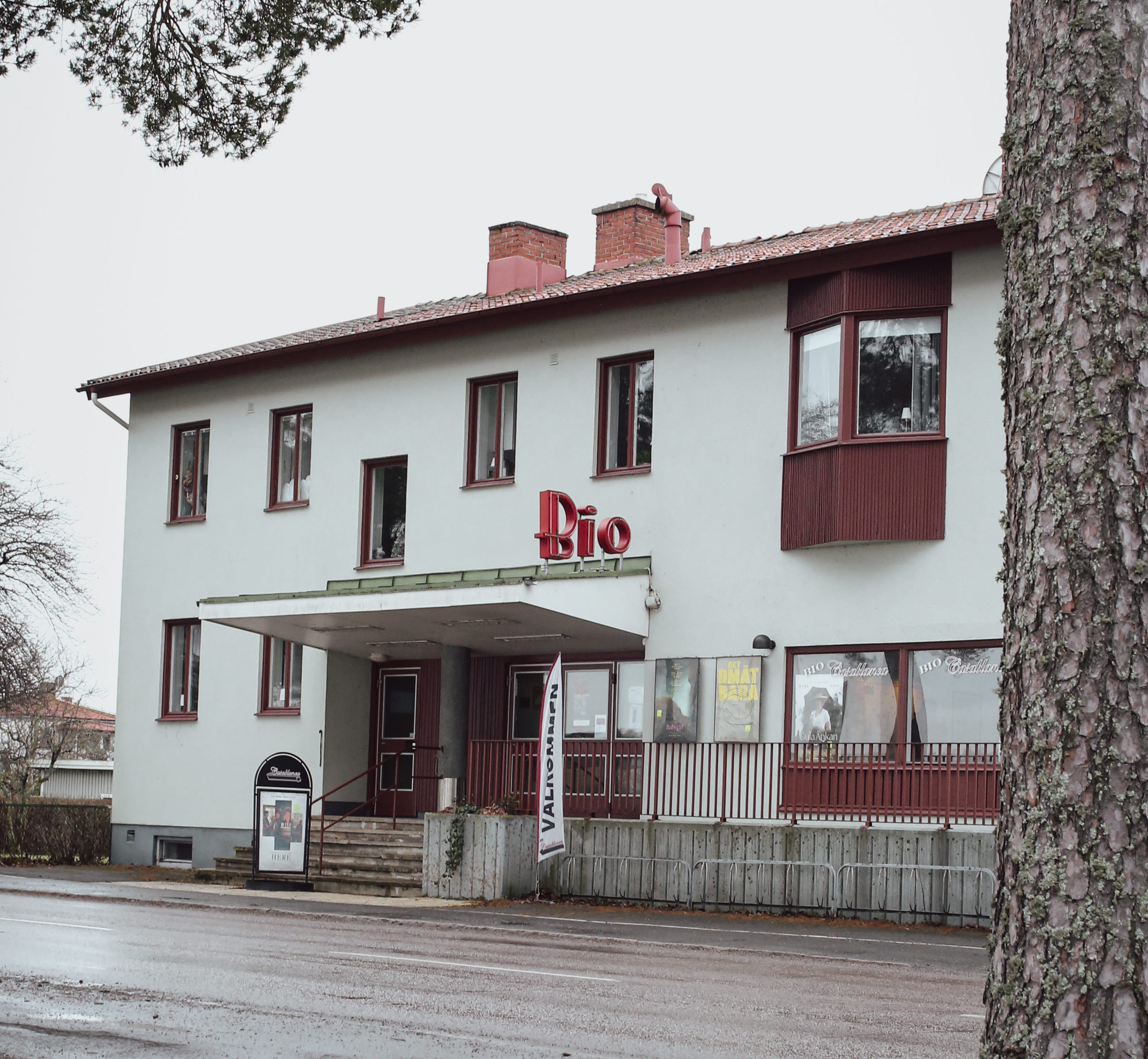 Ett foto av ett hyreshus med en biografentré centralt placerad på byggnadens framsida. I förgrunden står en hög tall som delvis skymmer byggnaden.