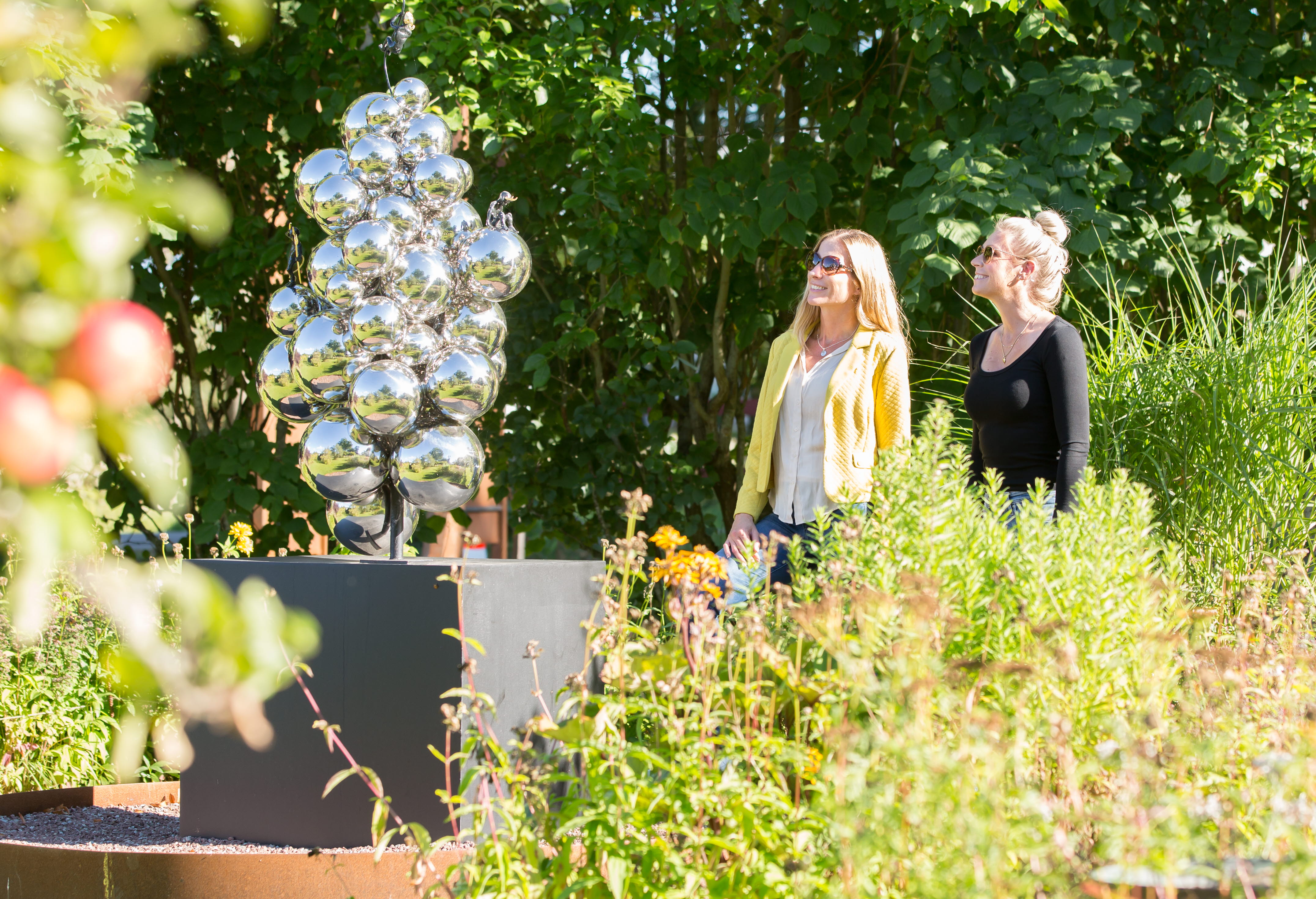 Två kvinnor som står och kollar på en skulptur med massa silvriga runda bubblor. Grönt runt om kring. 