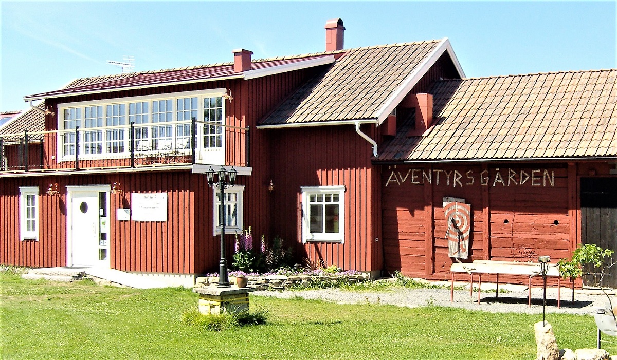 Röd träbyggnad med texten "Äventyrsgården" på väggen. Framför finns en gräsmatta med en stolpe mitt i.
