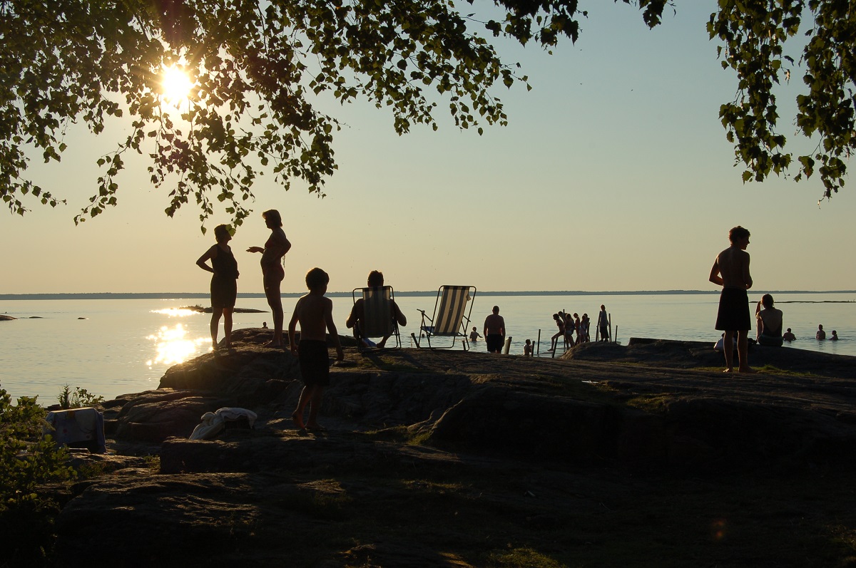 En vacker och stilla sommarkväll vid vattnet. I motljuset ser man människor och solstolar på klipporna och i förgrunden syns en björk. 