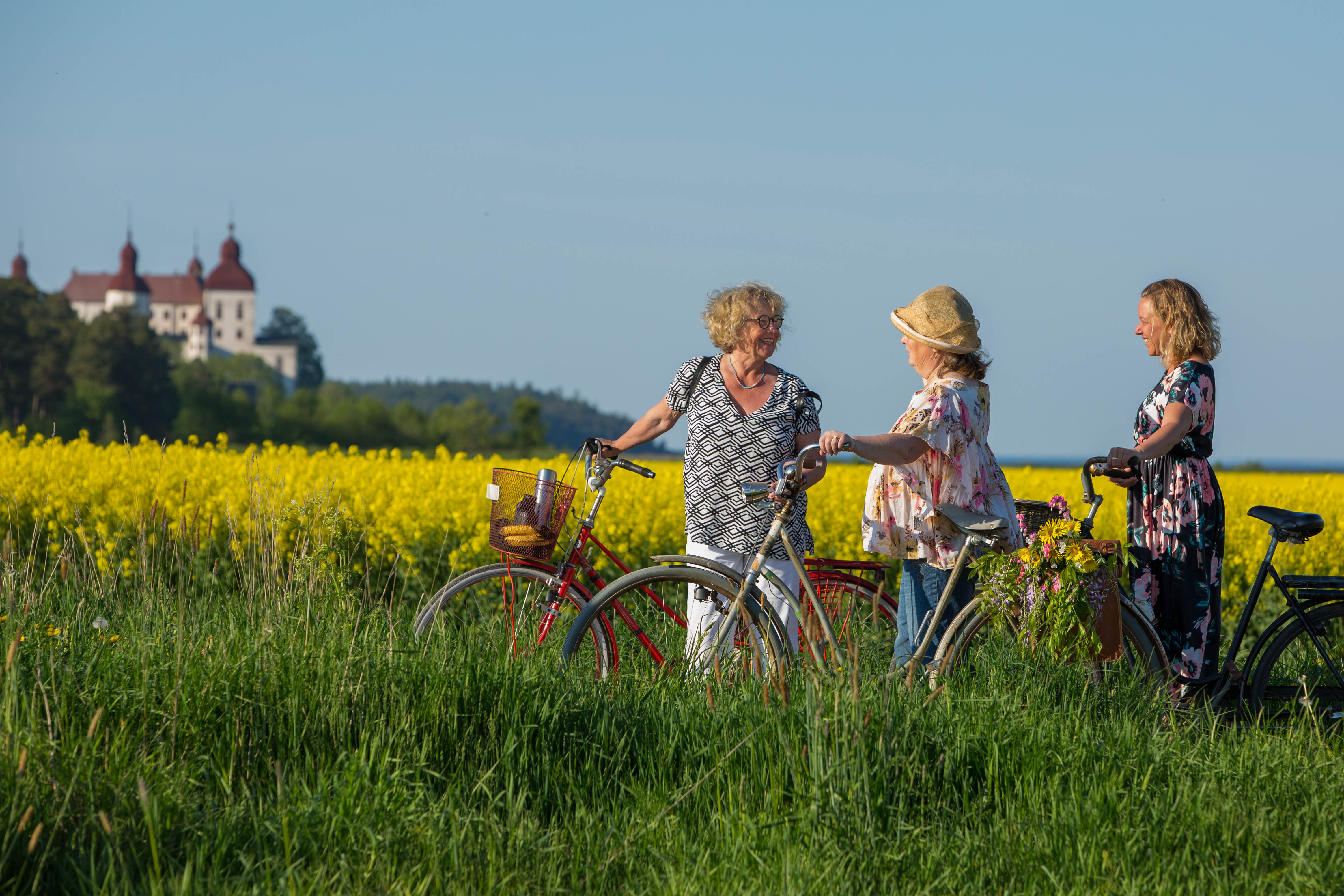 Tre kvinnor står med varsin cykel på ett fält med gula blommor. Bakom dem syns ett vitt slott (Läckö Slott). 