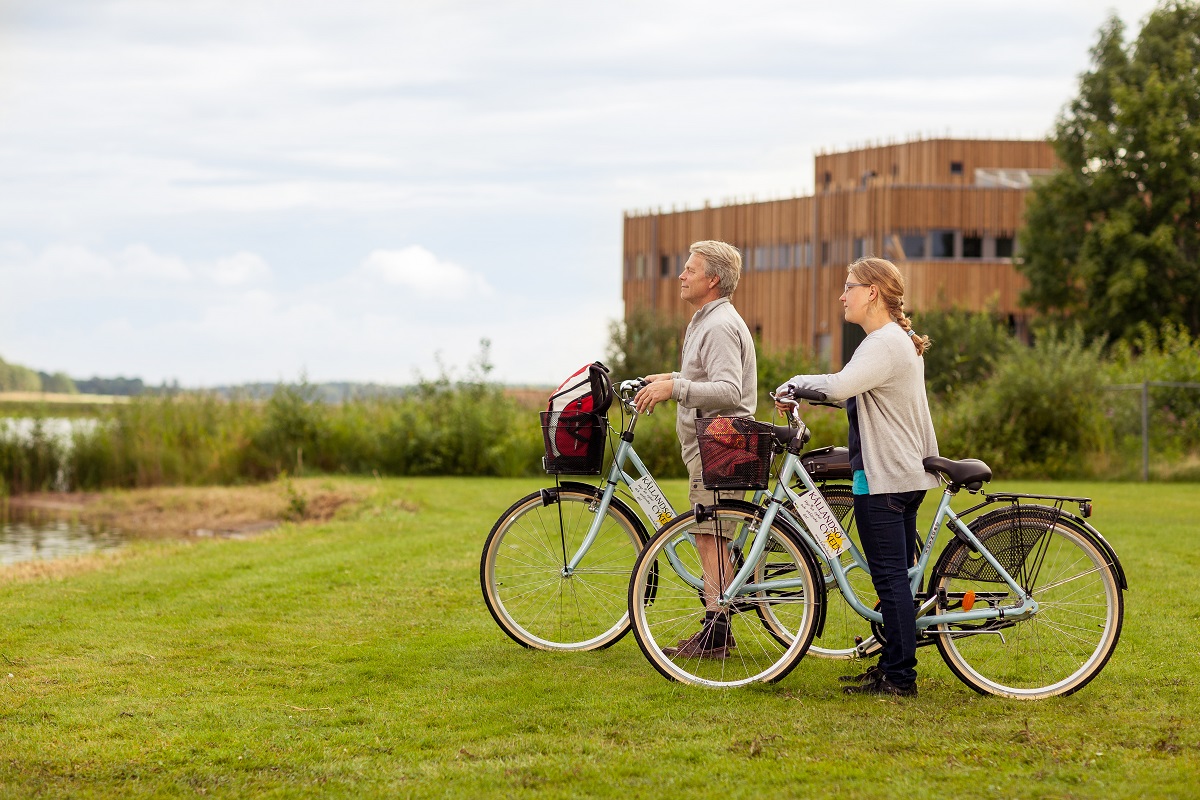 A man and a woman stand with their bikes on a green lawn one possible day. In the background is a brown wooden building.