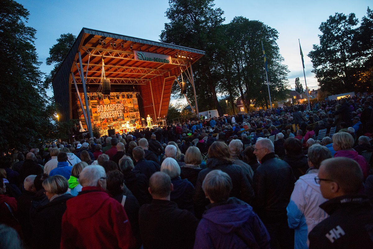Konsert på utomhusscen i Stadsträdgården i Lidköping. Scenen lyser upp och framför står ett hav av människor.