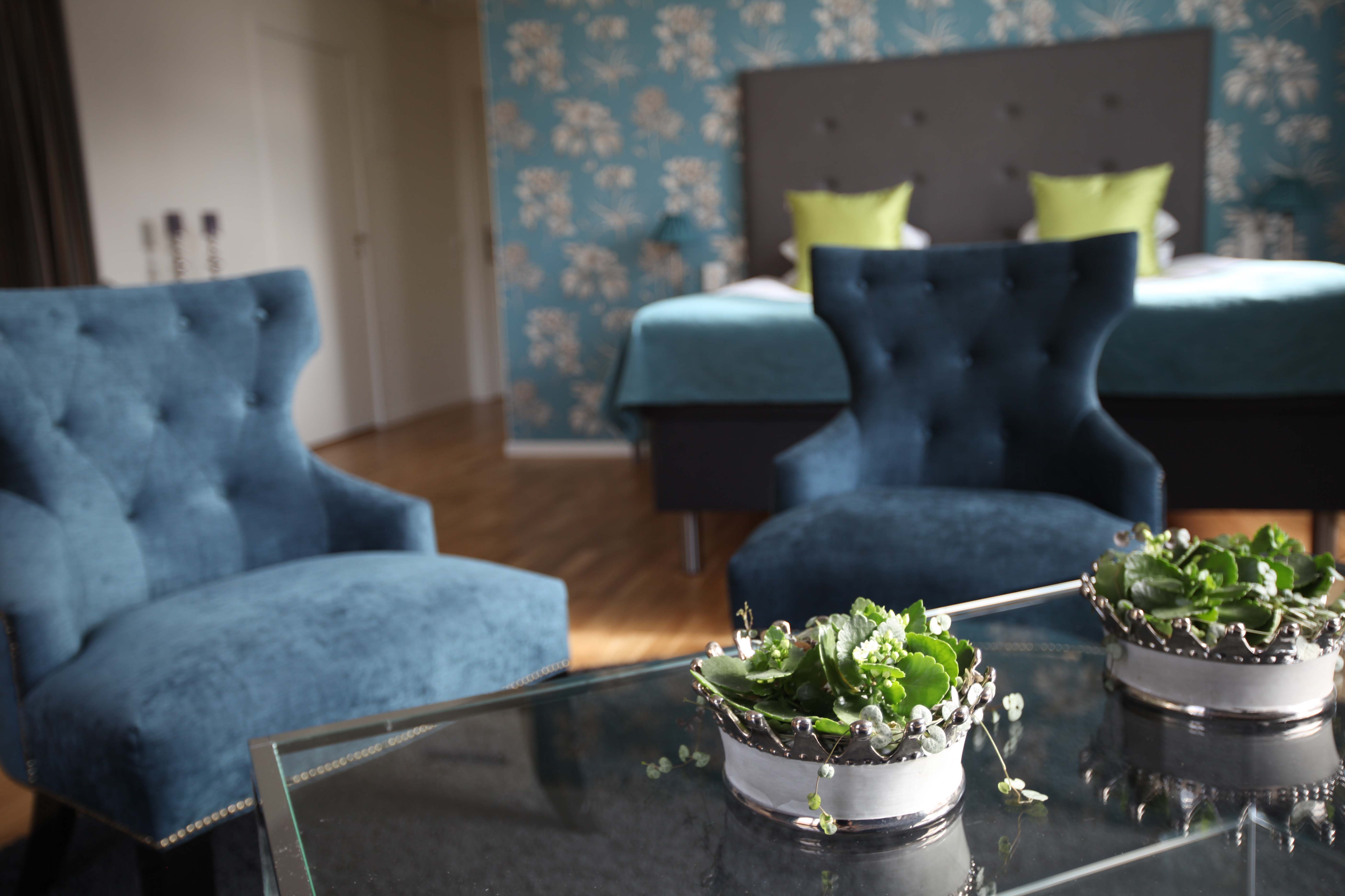 Hotellrum med en dubbelsäng i bakgrunden och två blå fåtöljer framför. I förgrunden finns ett bord med två blomkrukor.