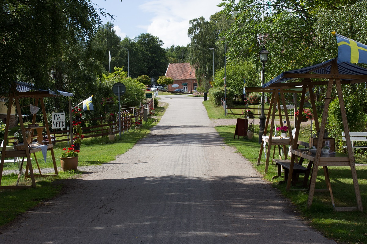 A wide gravelroad in Falkängens hantverksby. Along the road is small stalls and a swedish flag is waving in the right corner.
