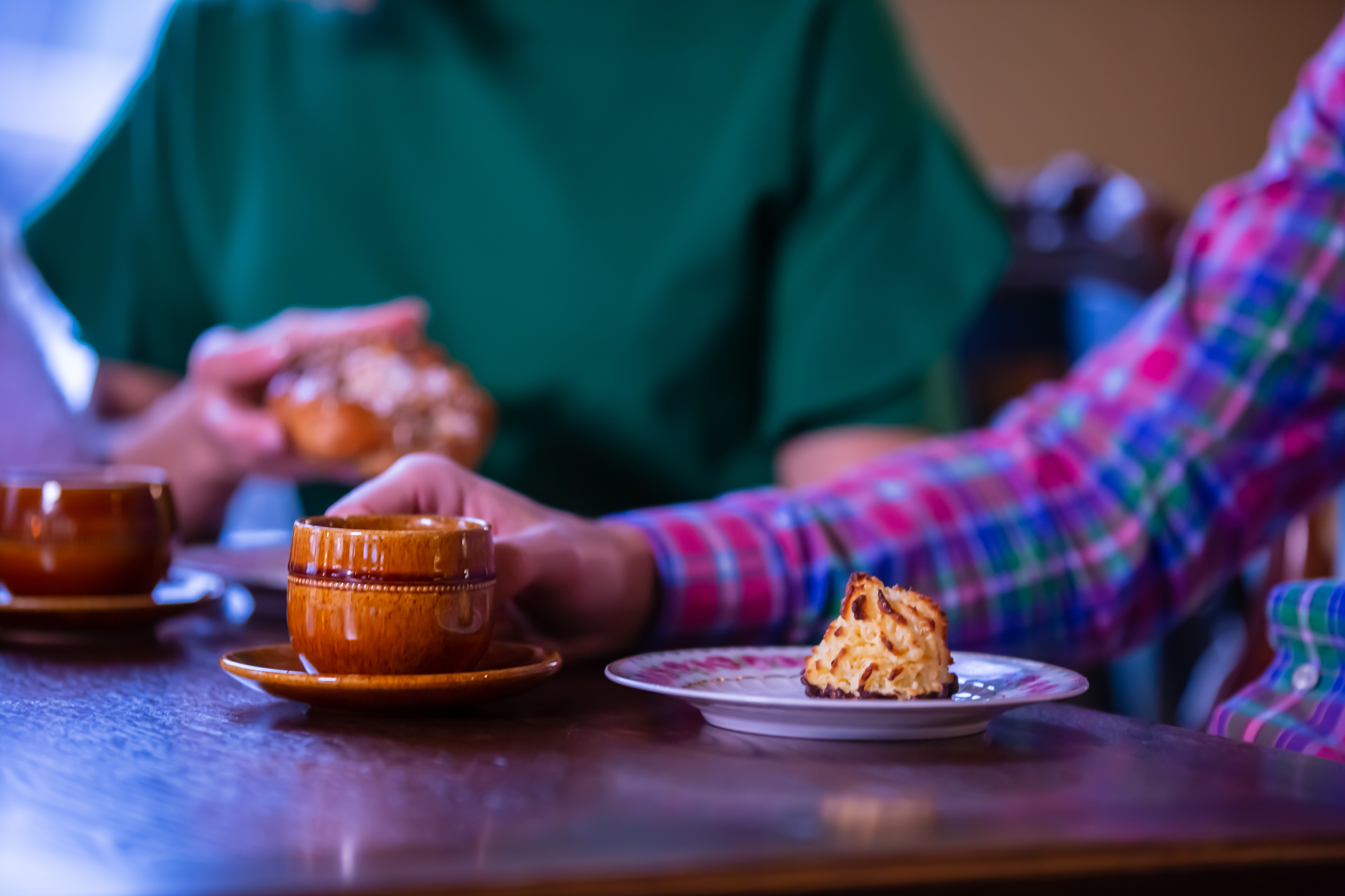 Två kaffekoppar och en kaka på en assiett. I bakgrunden syns en hand som håller i en bulle. 