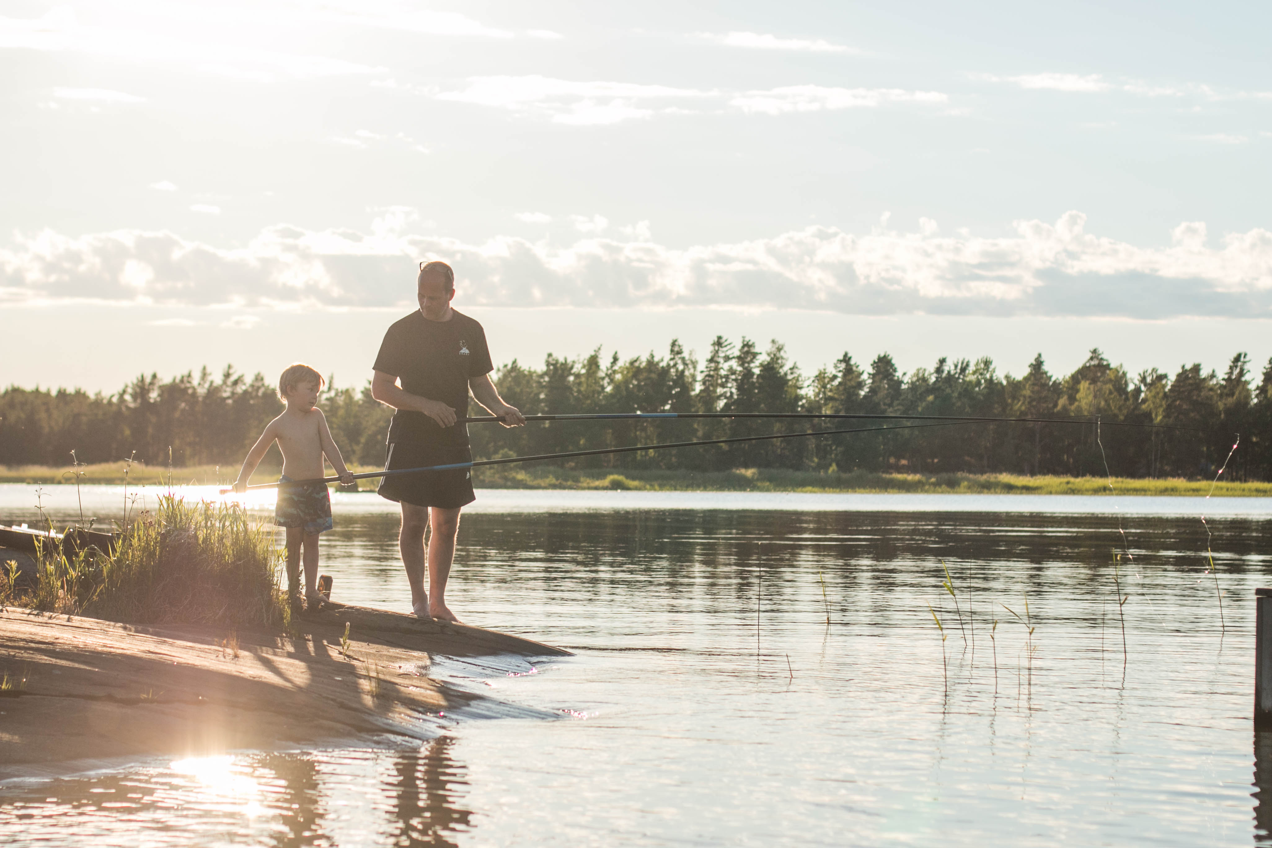 En vuxen och ett barn står med varsitt fiskespö i händerna vid vattnet. Bakom dem syns skog och en solnedgång. 