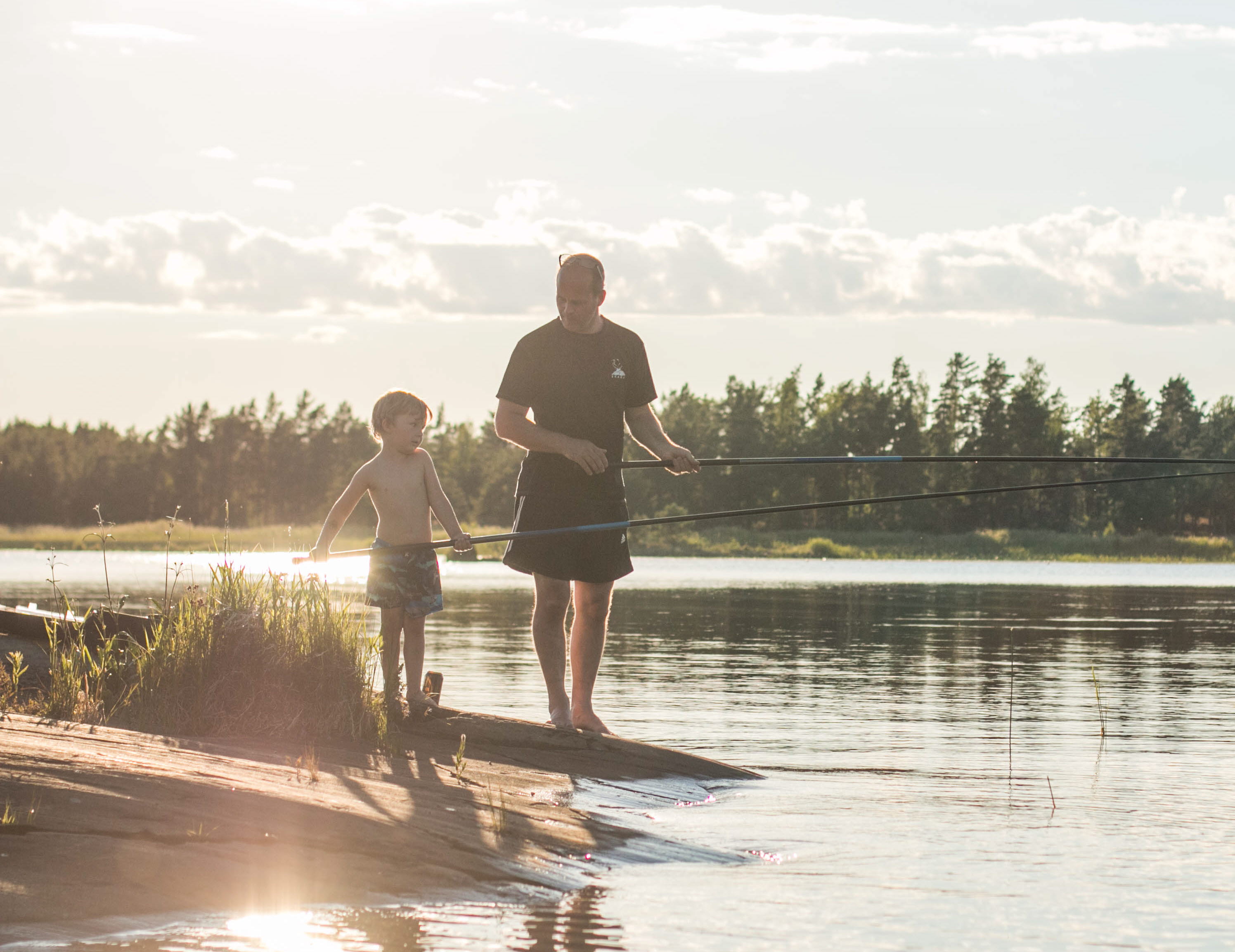 An adult and a child stand with their fishing rods in their hands by the water. Behind them you can see forest and a sunset.